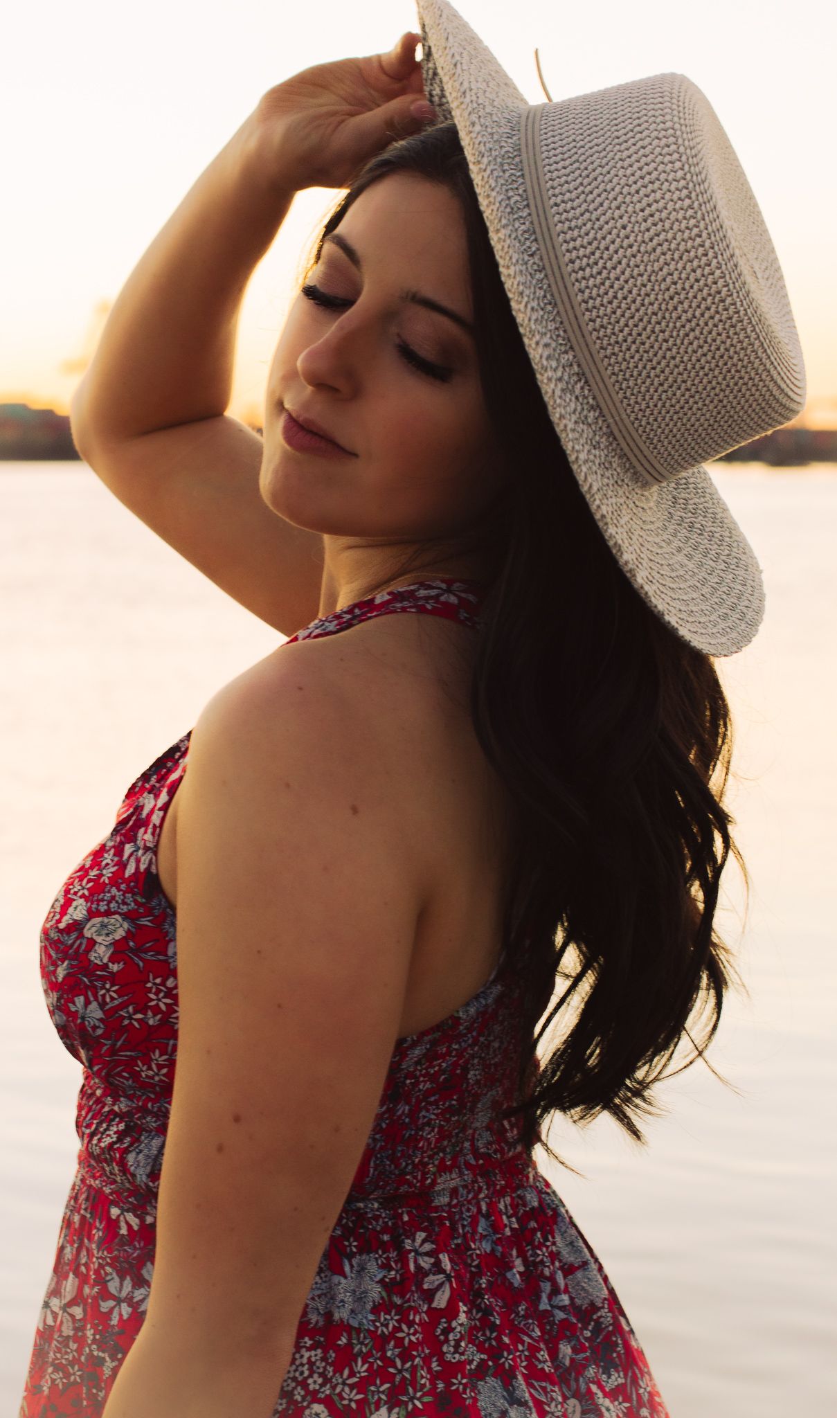 Woman in red floral dress and white sun hat poses during sunset beach photoshoot.