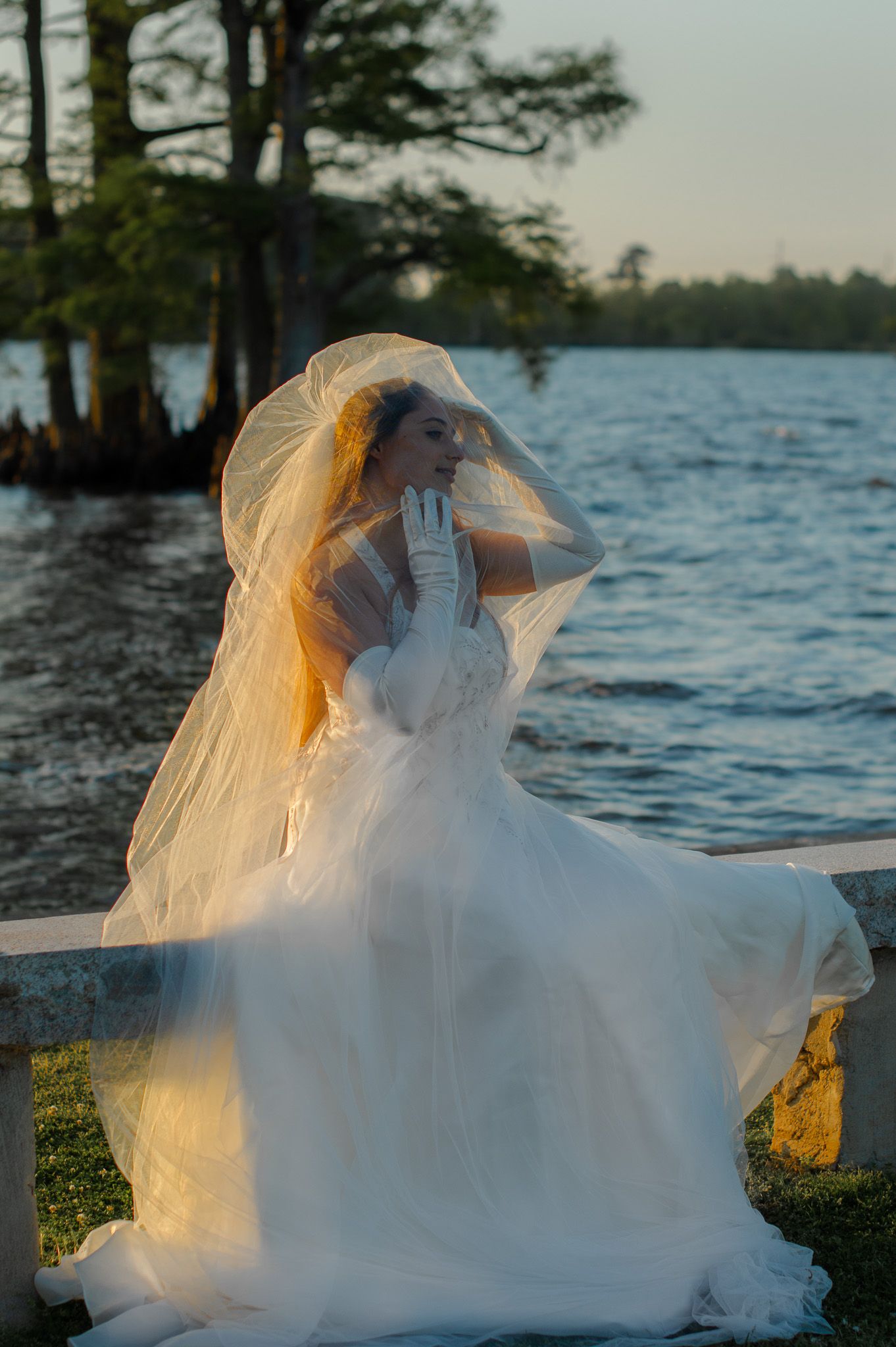 Bride in white gown and veil sits by a serene lake at sunset, gloves on hands, dress flowing gracefully in the breeze.