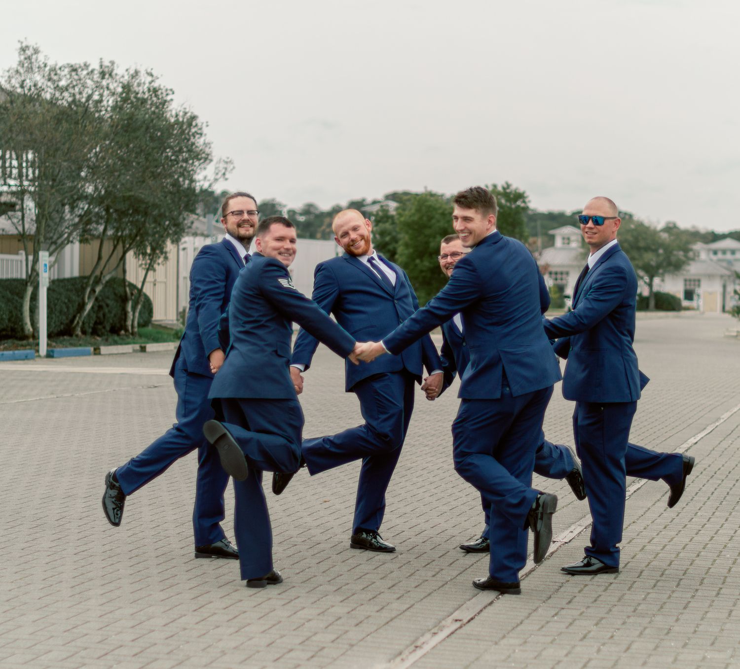 Groomsmen in navy suits perform synchronized kicks in a fun outdoor photo.