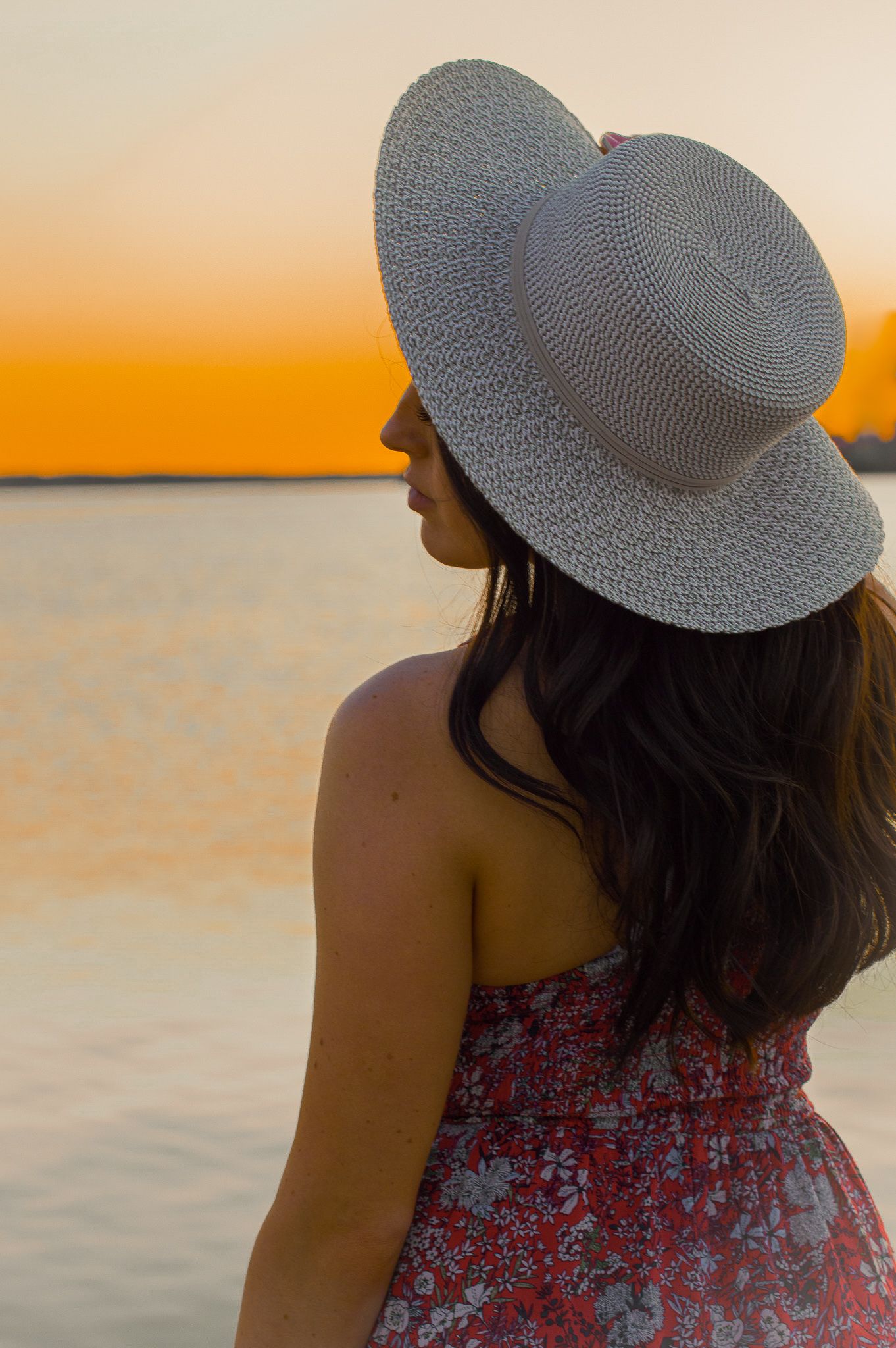 Person wearing white sun hat and floral dress gazes at sunset over water.