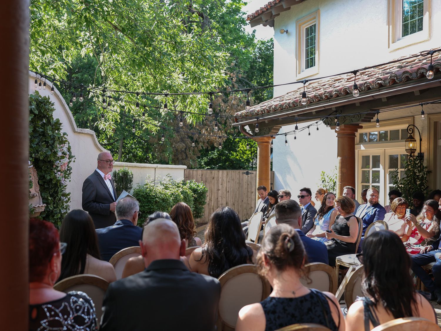 Outdoor wedding ceremony in a courtyard with guests seated facing a speaker near a Spanish-style building surrounded by trees.