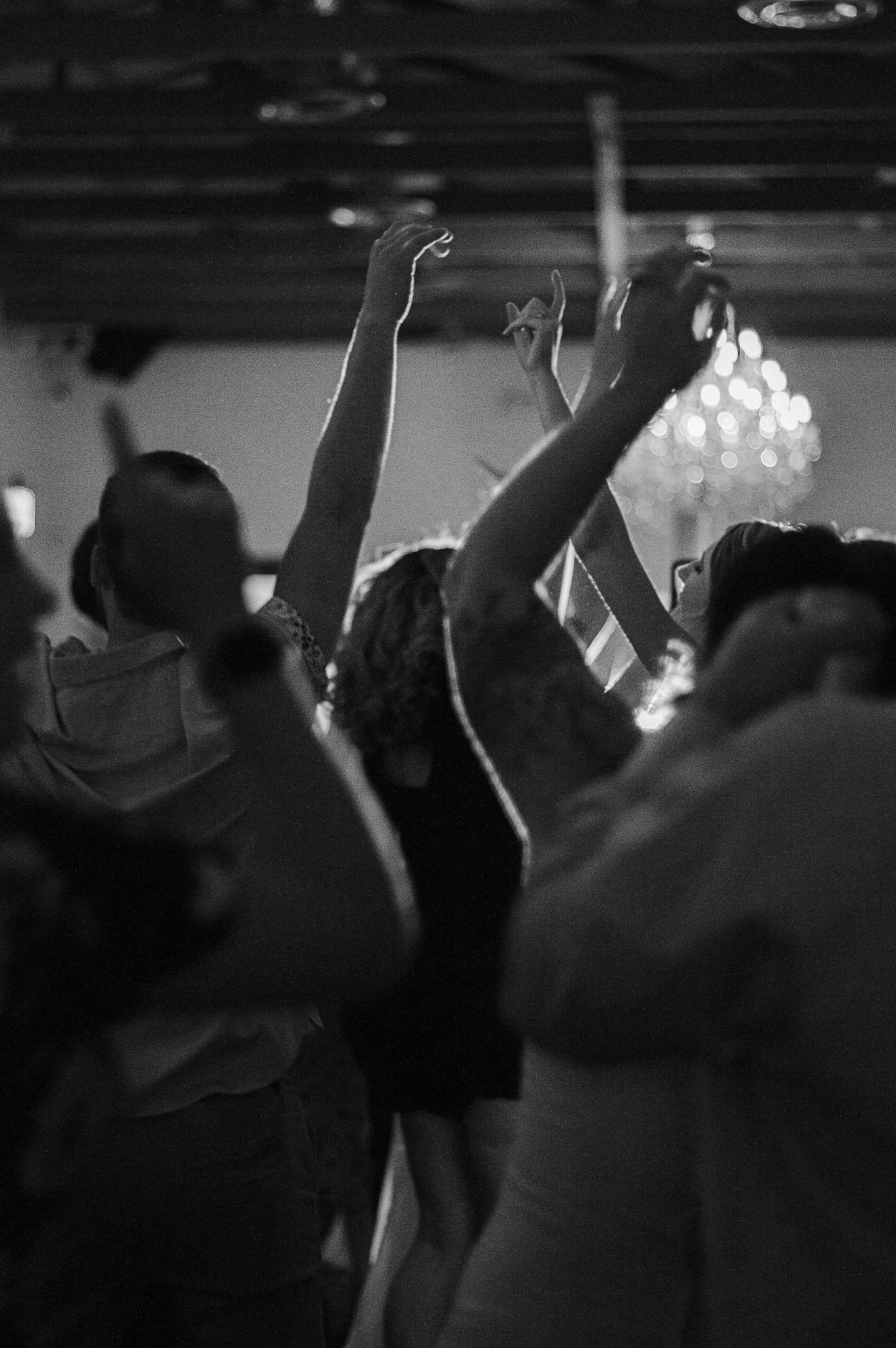 Crowd of party guests dancing with raised arms under dim chandelier lighting.