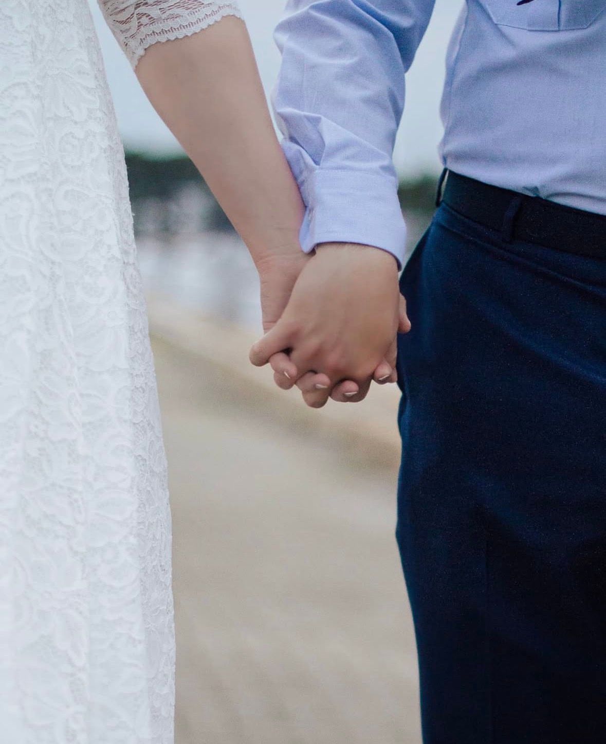A close-up view of newlyweds holding hands, with the bride in white lace and groom in blue dress shirt and navy pants.