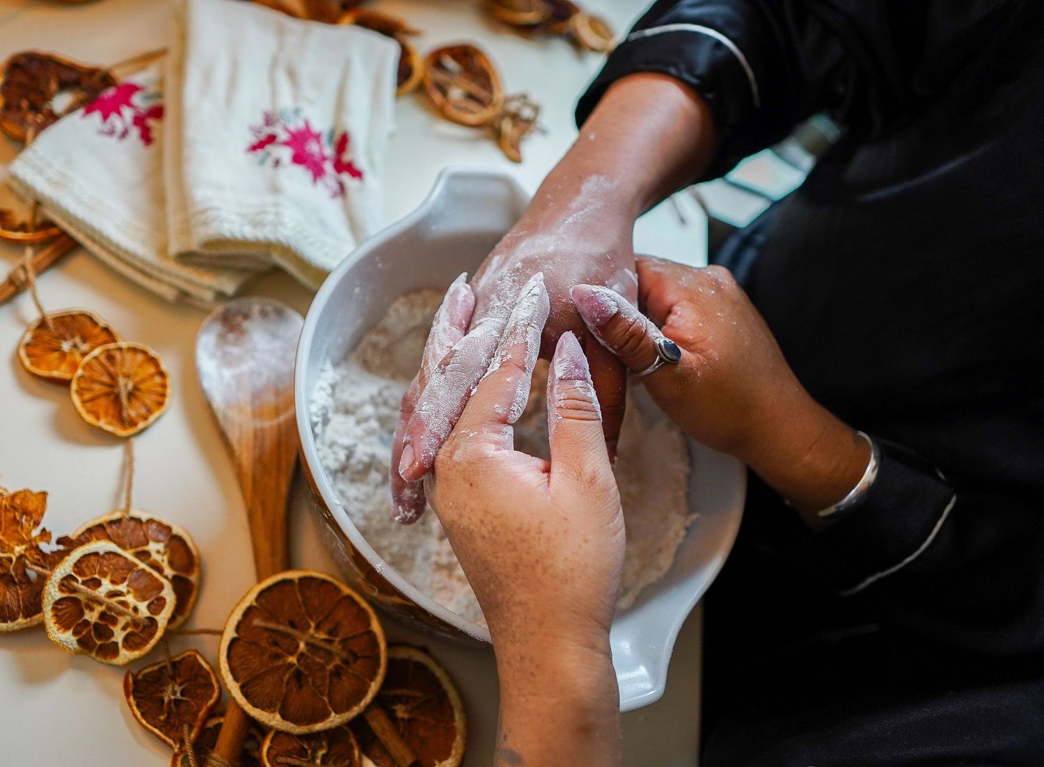 Two hands being washed with flour in a white bowl, surrounded by dried orange slices and linen napkins.