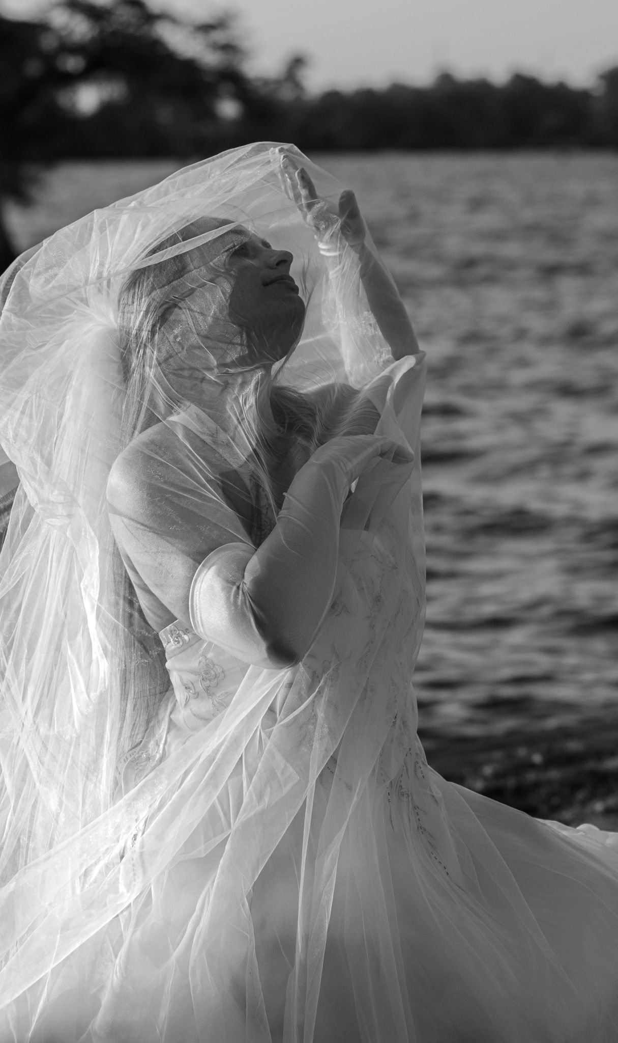 Black and white photo of a bride in a flowing white gown and veil posing gracefully by a serene lake.
