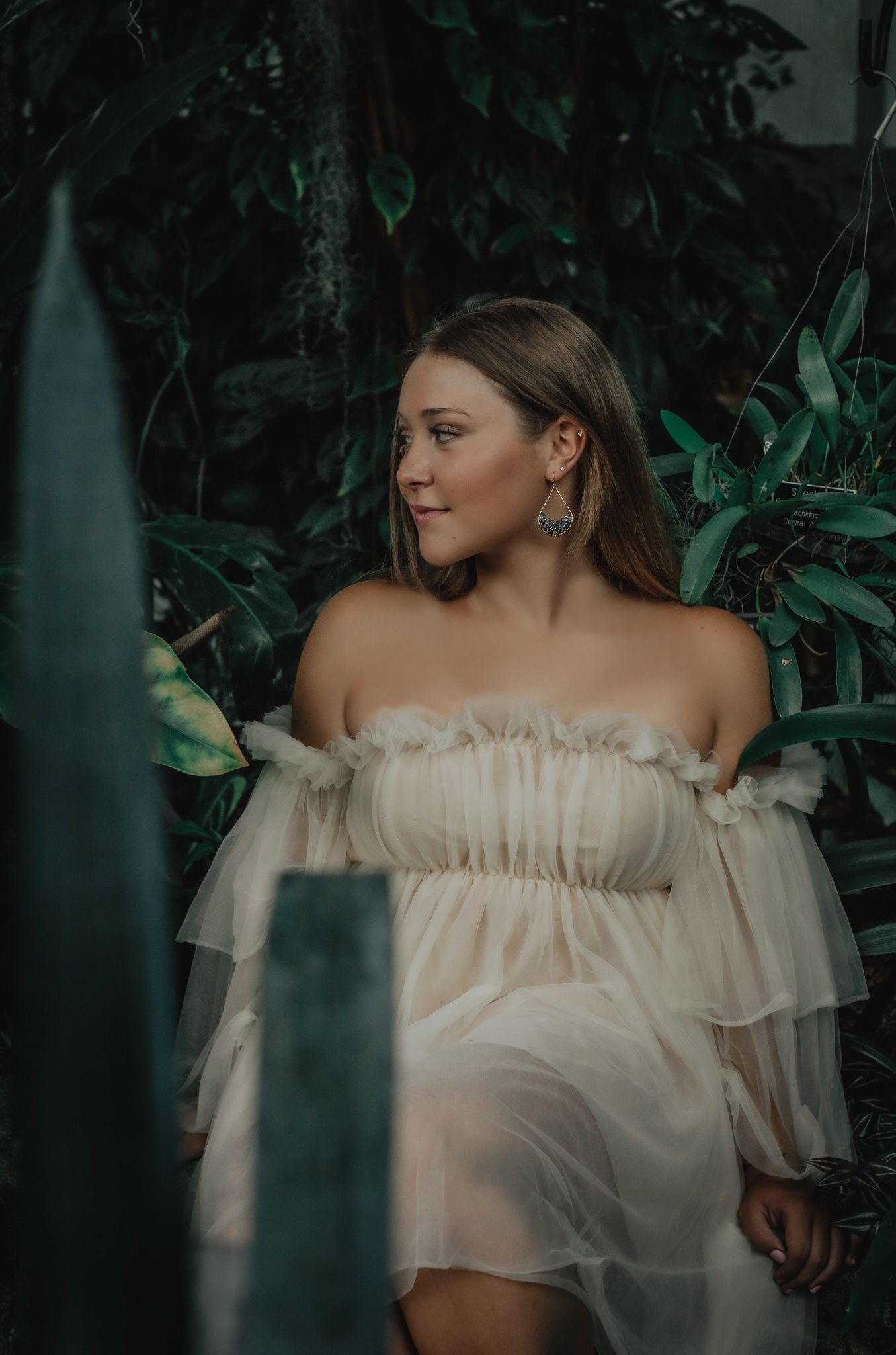 Ethereal portrait in white off-shoulder dress against dark leafy background.