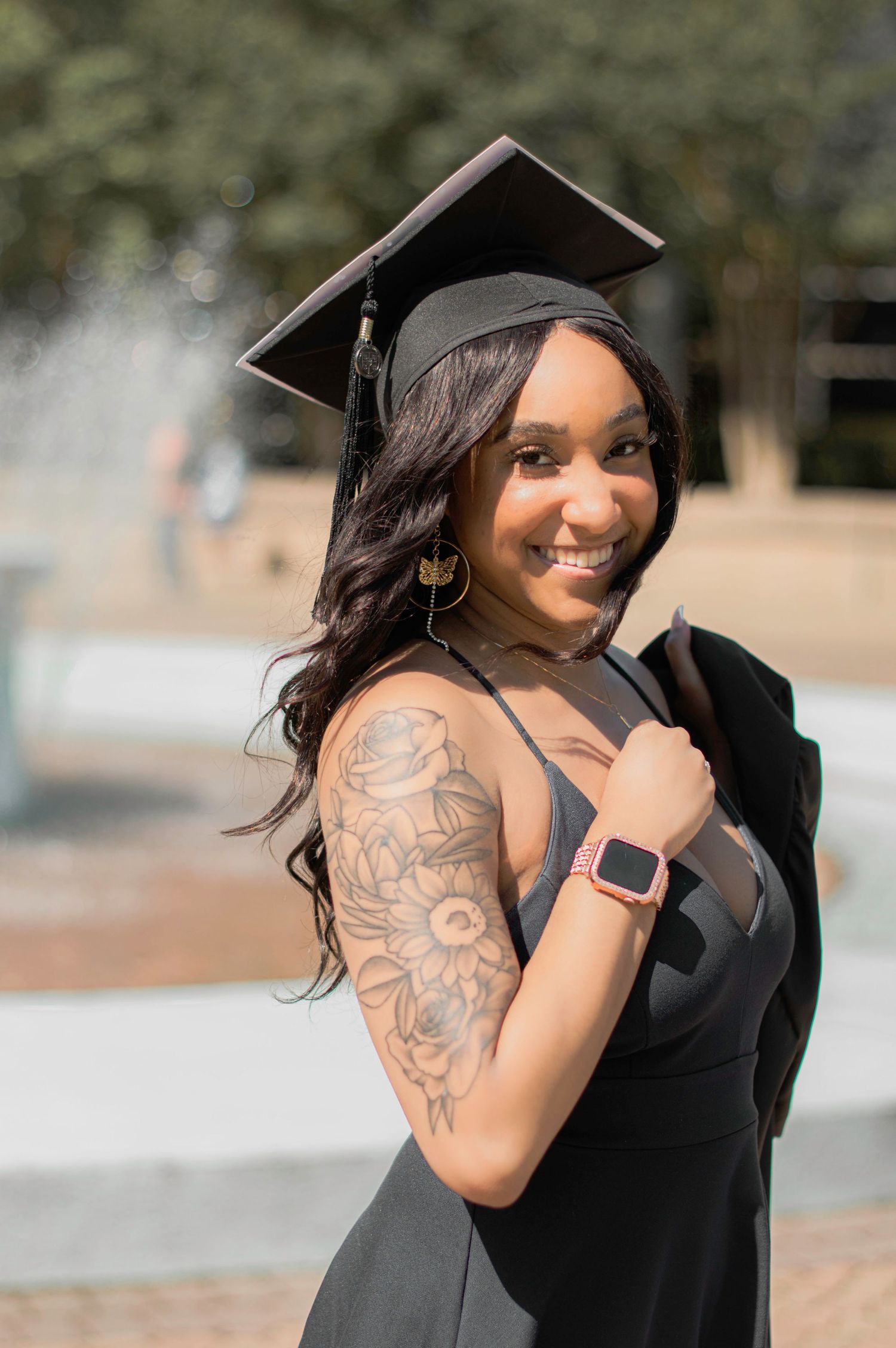 Graduate in black cap and gown shows off arm tattoo while posing outdoors.
