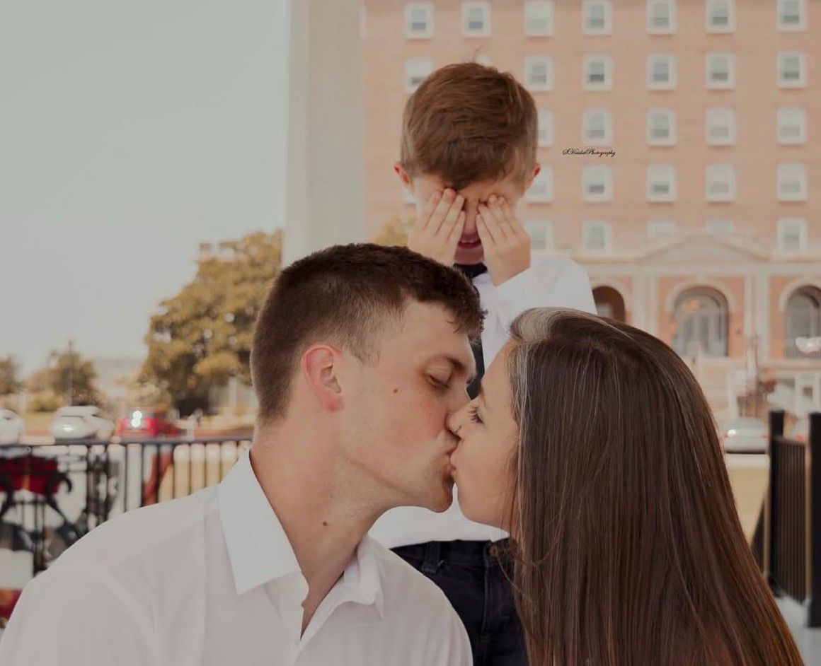 A romantic kiss shared between a couple on a sunny day with a brick building in the background.
