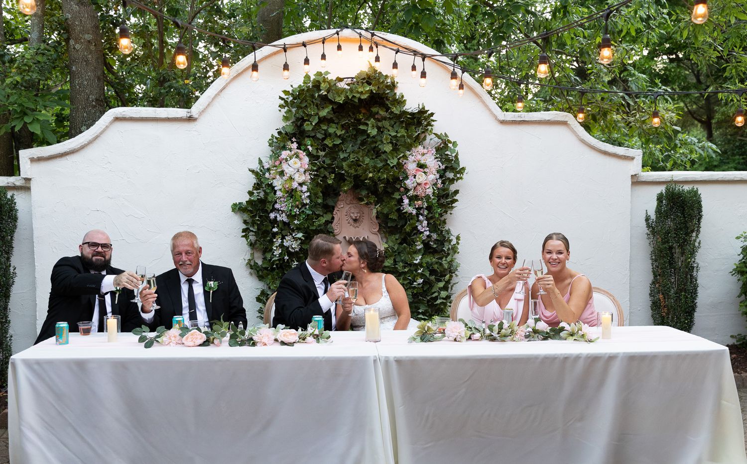Wedding party laughing at sweetheart table with floral arch backdrop and string lights at outdoor reception.