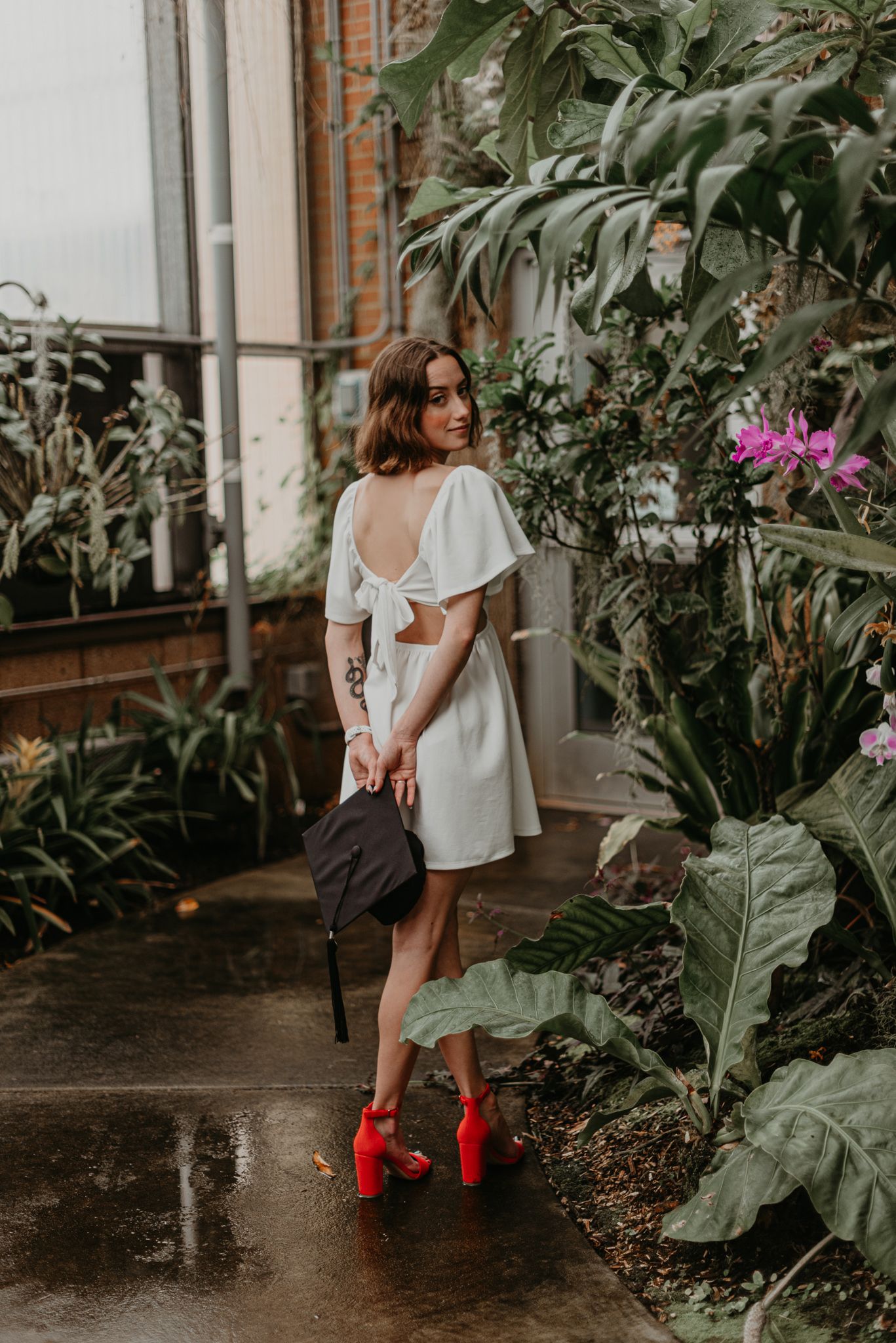 Graduate in white dress and red boots poses among greenhouse plants.
