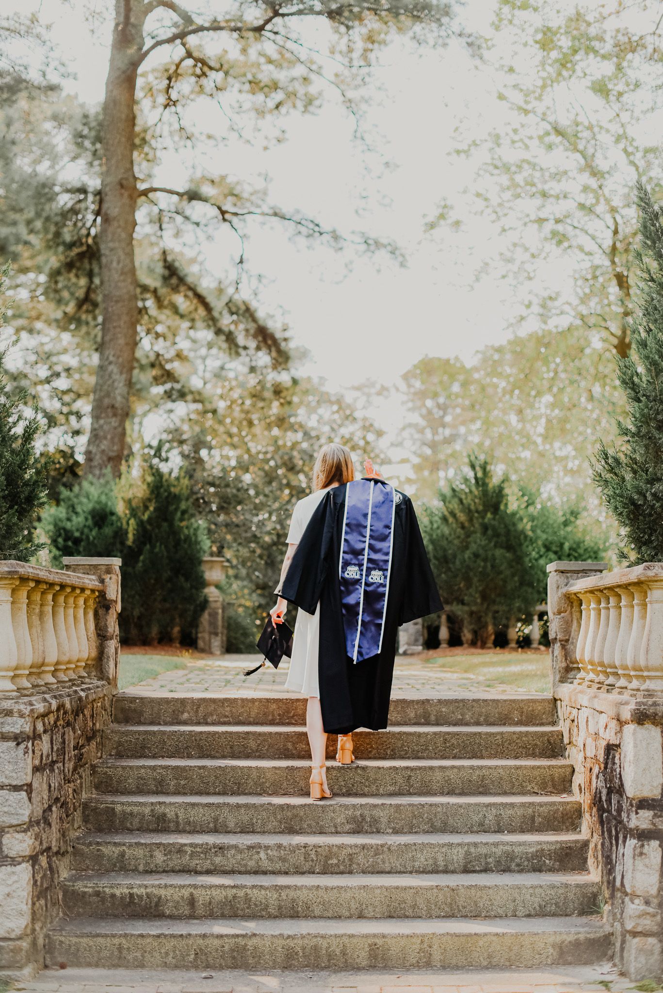 A graduate ascending stone steps in cap and gown surrounded by trees.