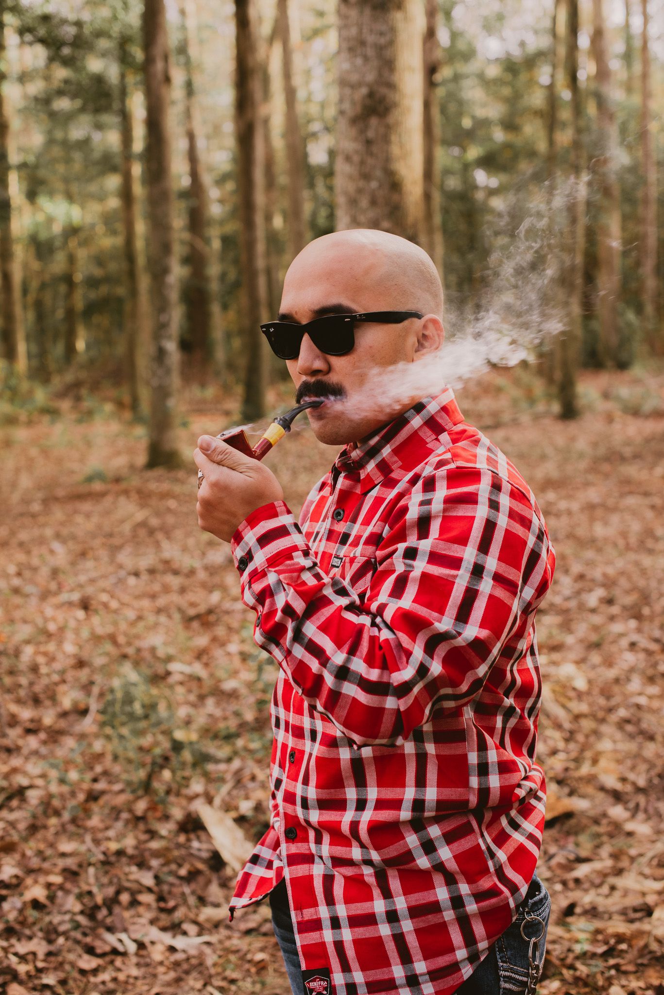 Person in red plaid flannel shirt and sunglasses smoking in autumn forest setting.