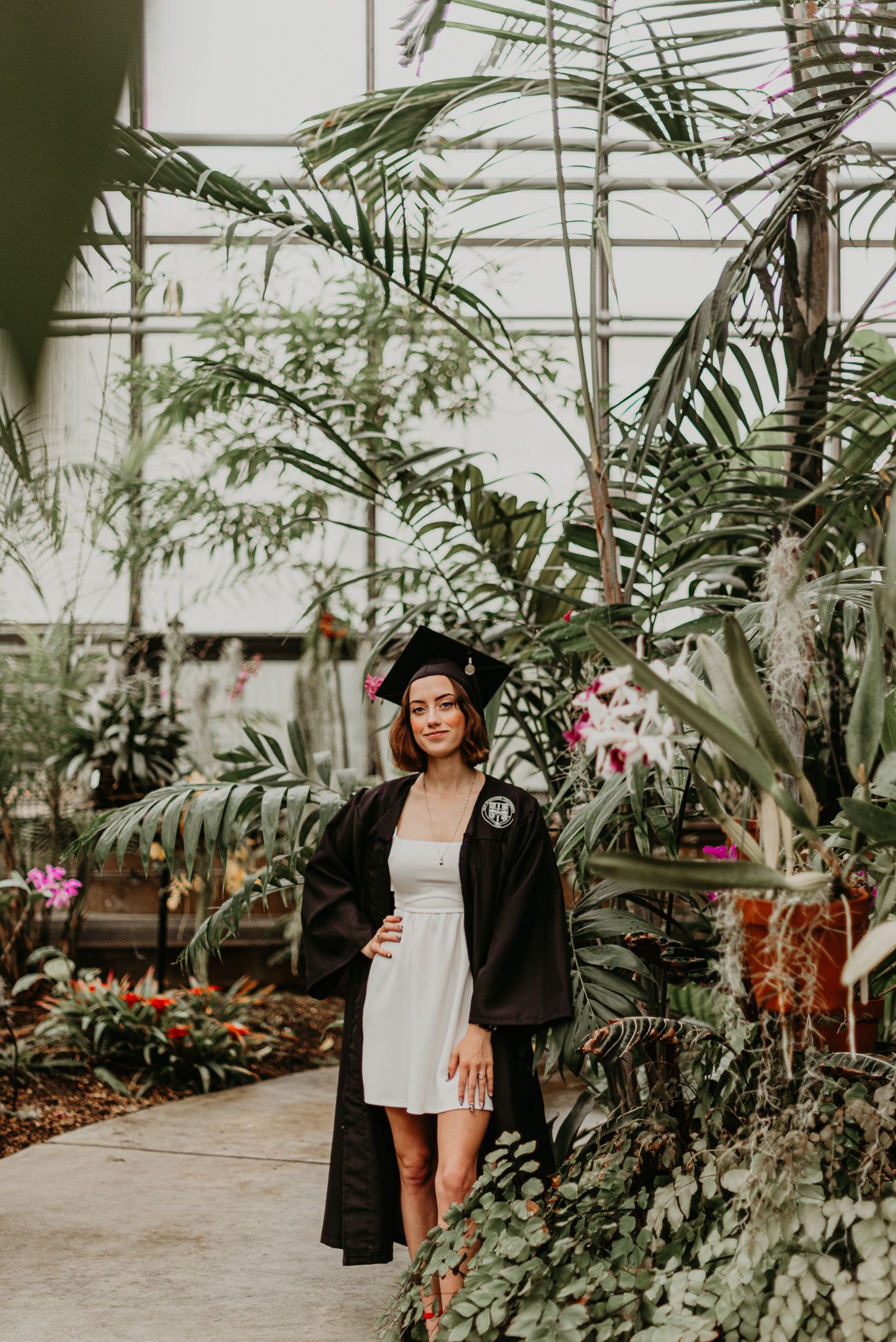 Graduate poses in white dress and black gown inside greenhouse with tropical plants.