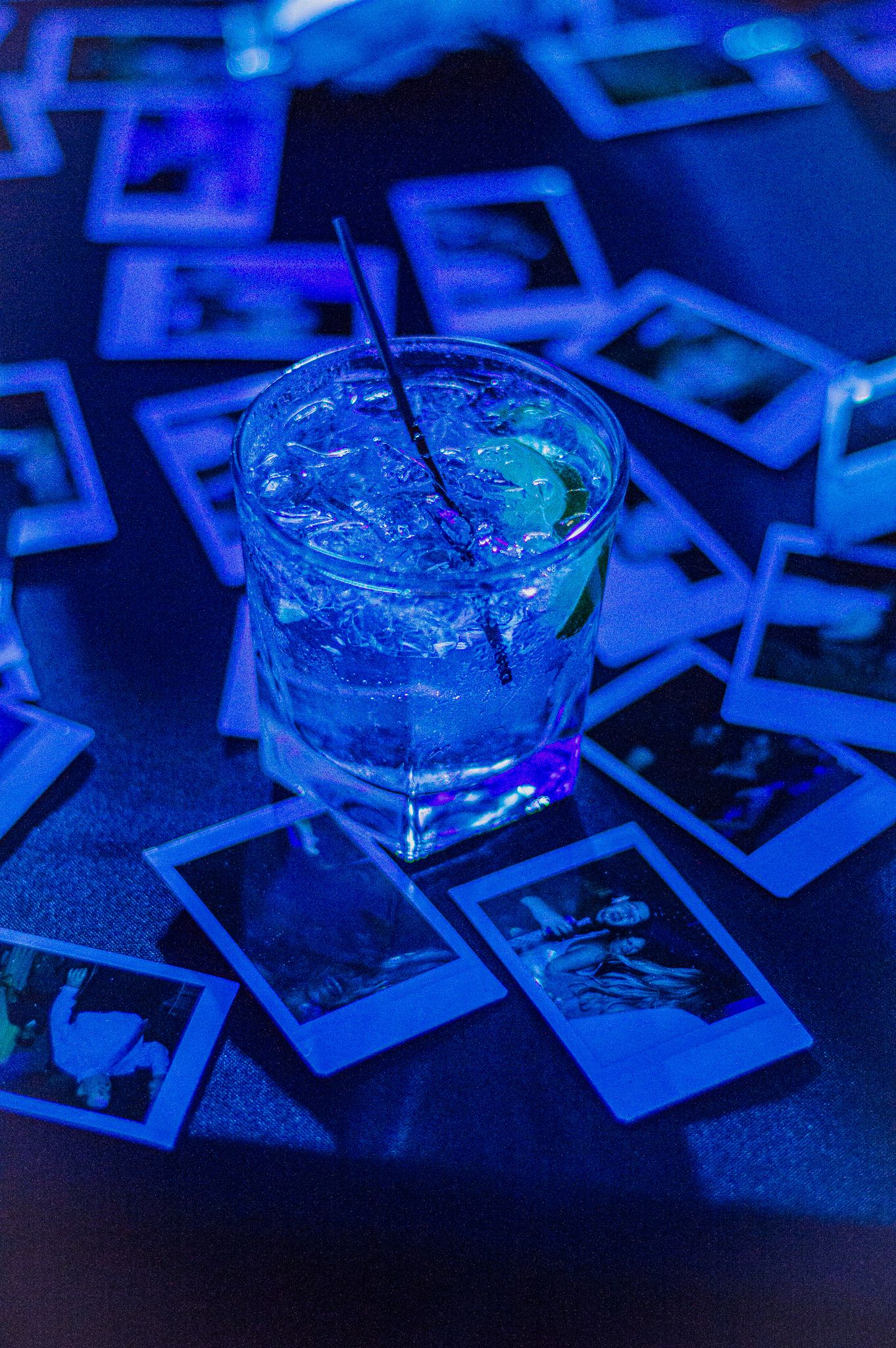 Blue cocktails on scattered polaroid photos under dramatic lighting.