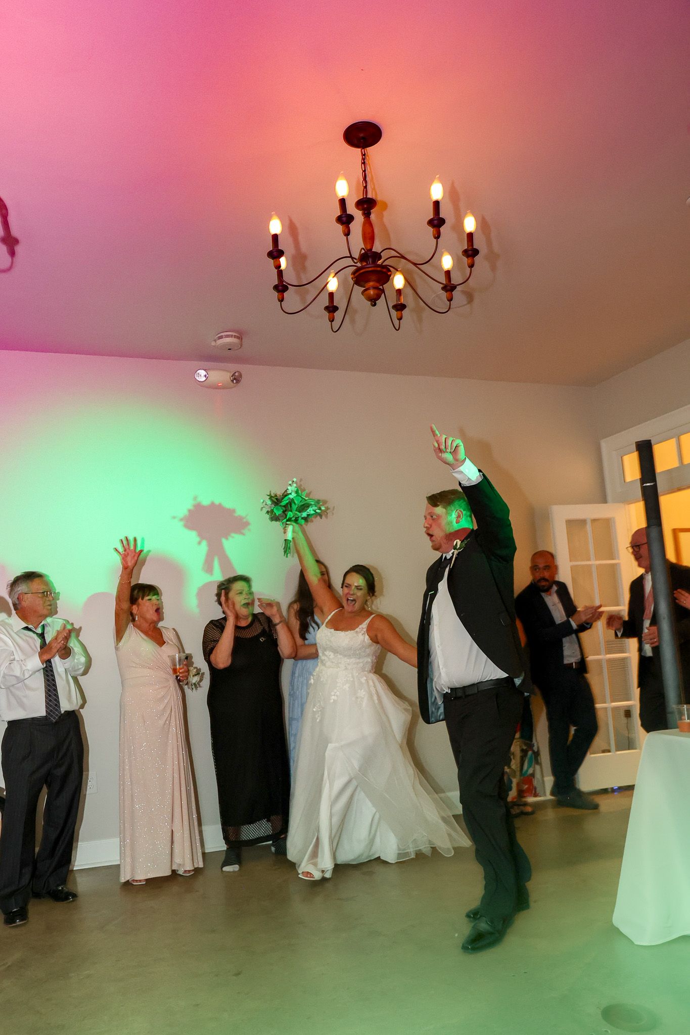 Bride and groom tossing bouquet at wedding reception with guests celebrating under chandelier with colorful pink and green lights.