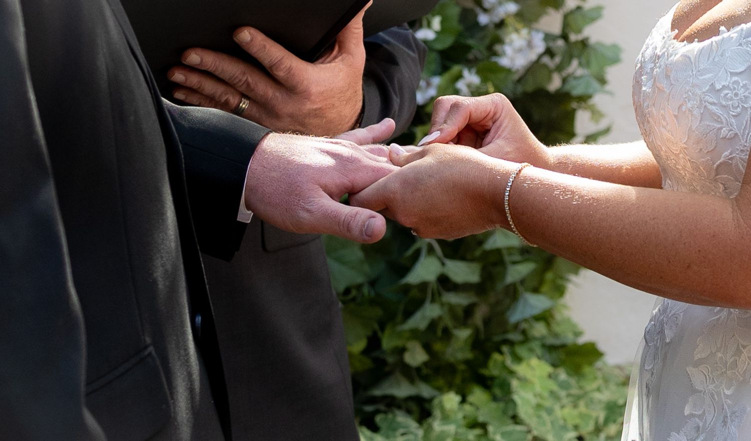 Bride placing wedding ring on groom's finger during outdoor ceremony with green floral backdrop.