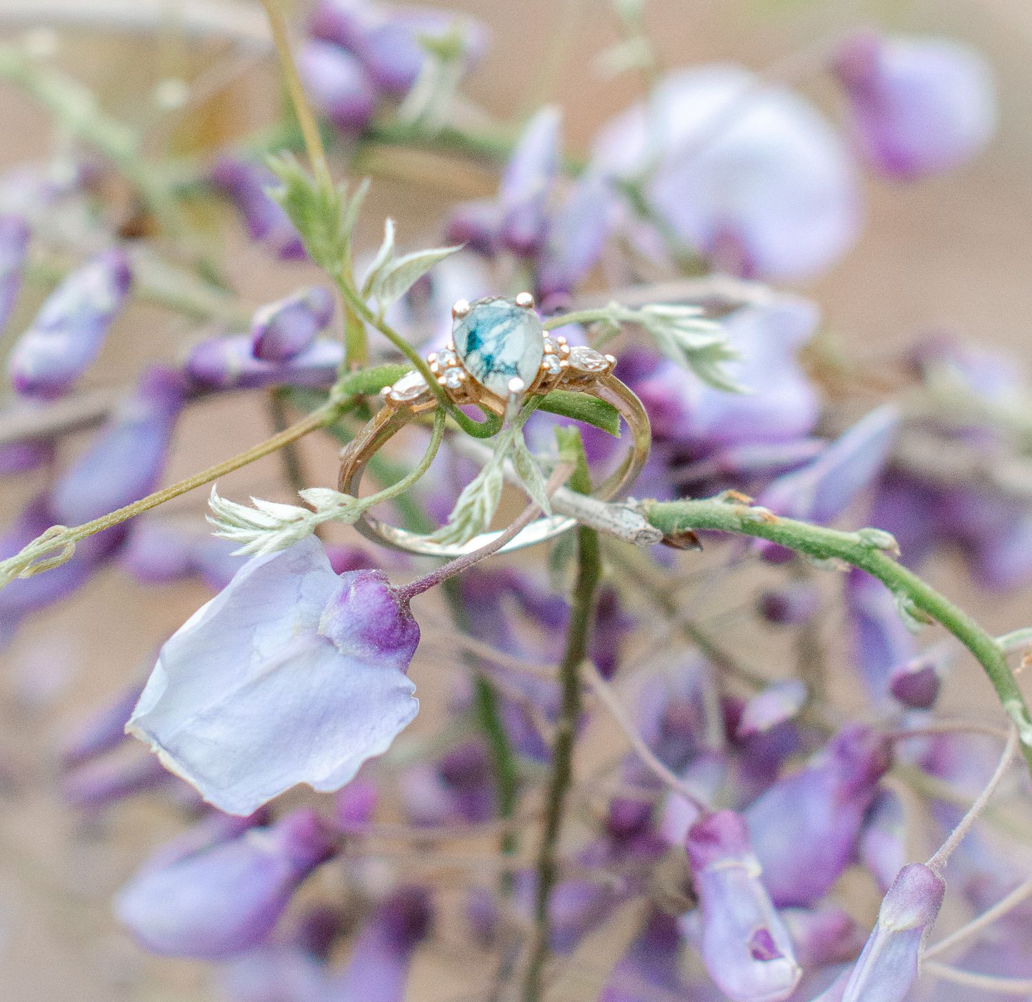 Gold engagement ring with aquamarine gemstone resting on purple wisteria flowers in soft bokeh background.