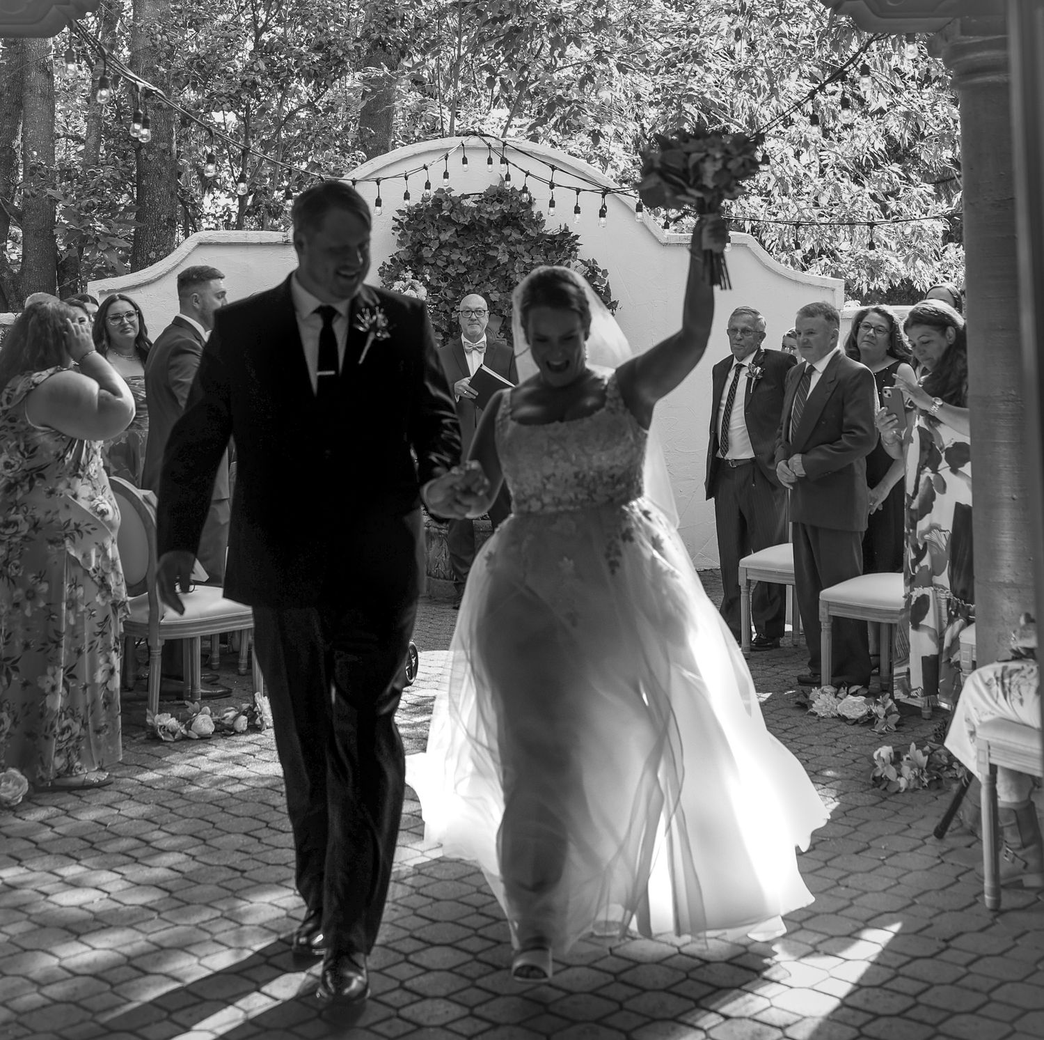 Bride raises bouquet triumphantly as couple walks back down the aisle after wedding ceremony outdoors.
