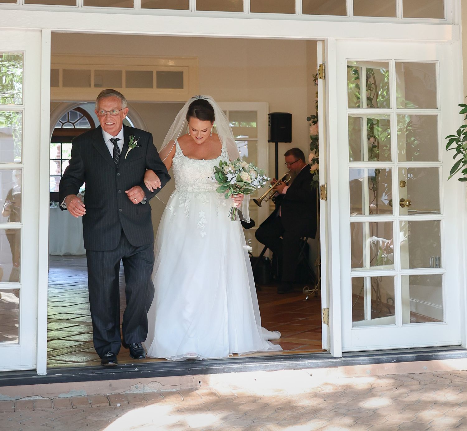 Bride in white gown walks through French doors with escort at outdoor wedding ceremony.