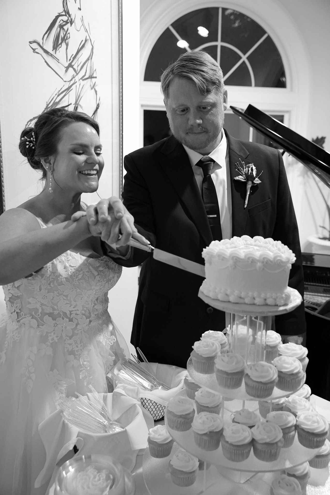 Bride and groom cutting wedding cake with cupcake tower at reception in black and white photo.