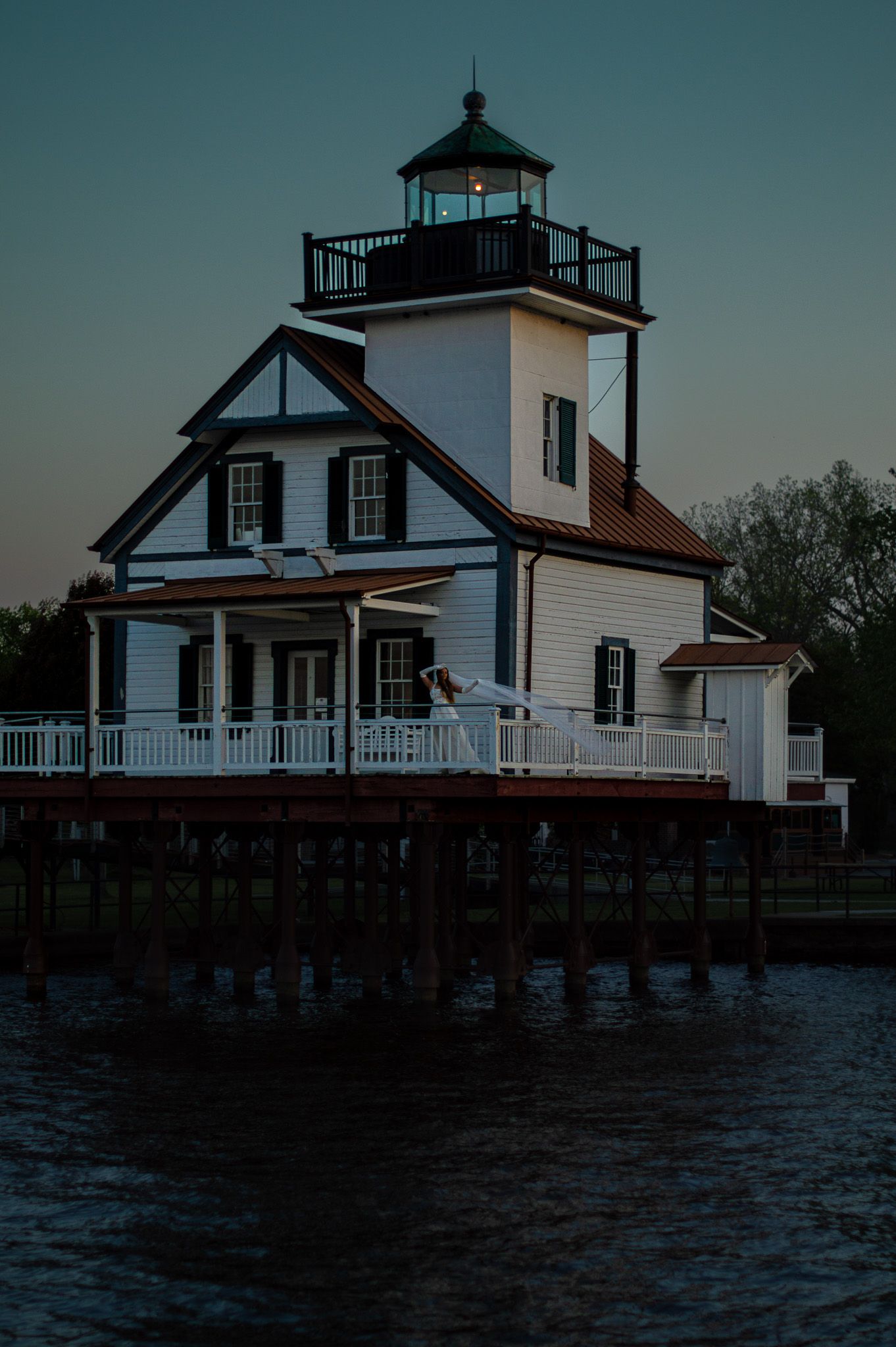 White lighthouse on stilts over water at dusk with glowing light, featuring wraparound porch and dark trim details.
