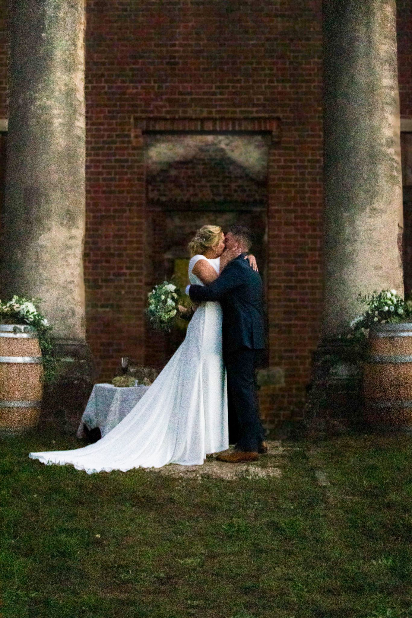 A romantic wedding couple shares an intimate moment between elegant stone columns against a brick building exterior.