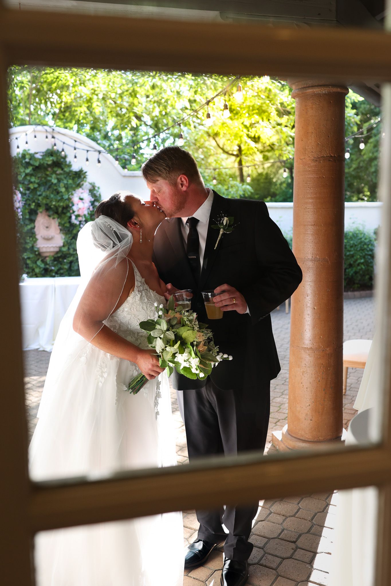 Bride and groom share a kiss at outdoor wedding ceremony, bride holding white floral bouquet, groom in black suit.