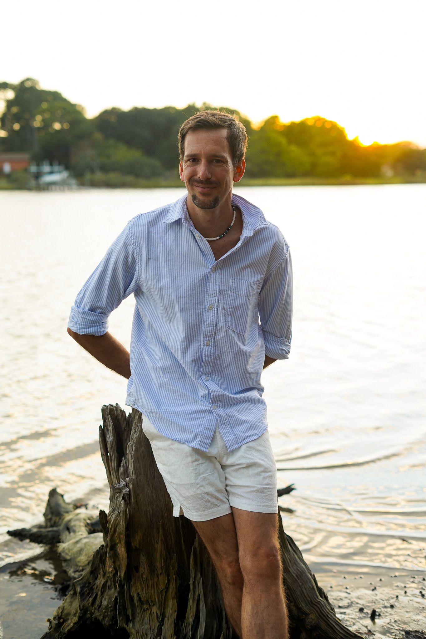 Person in white shirt sitting on driftwood at sunset by a tranquil beach.