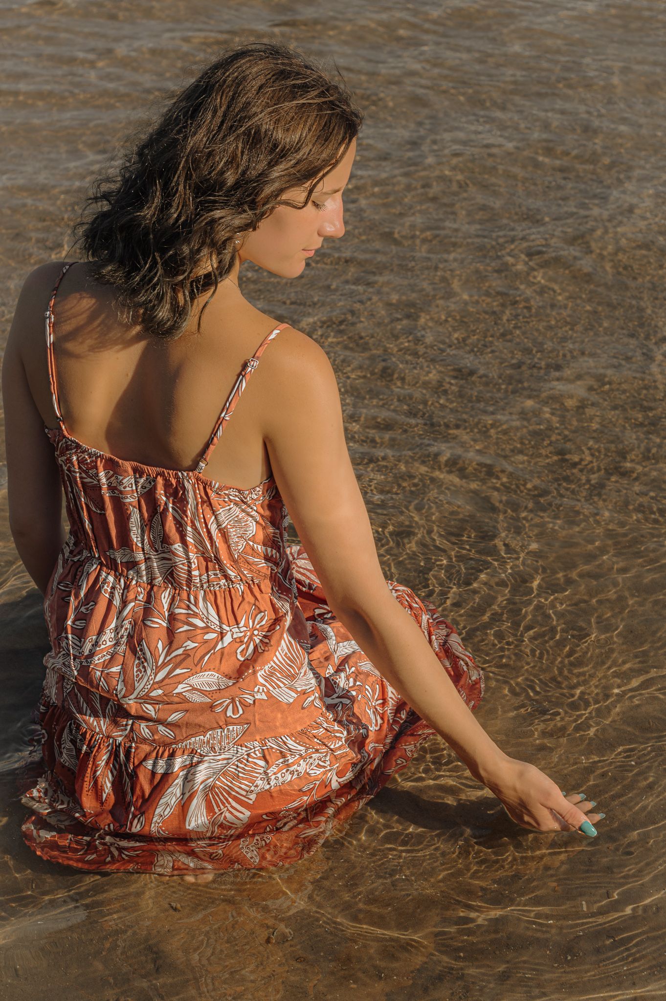 Person in floral sundress sits at water's edge during golden hour.