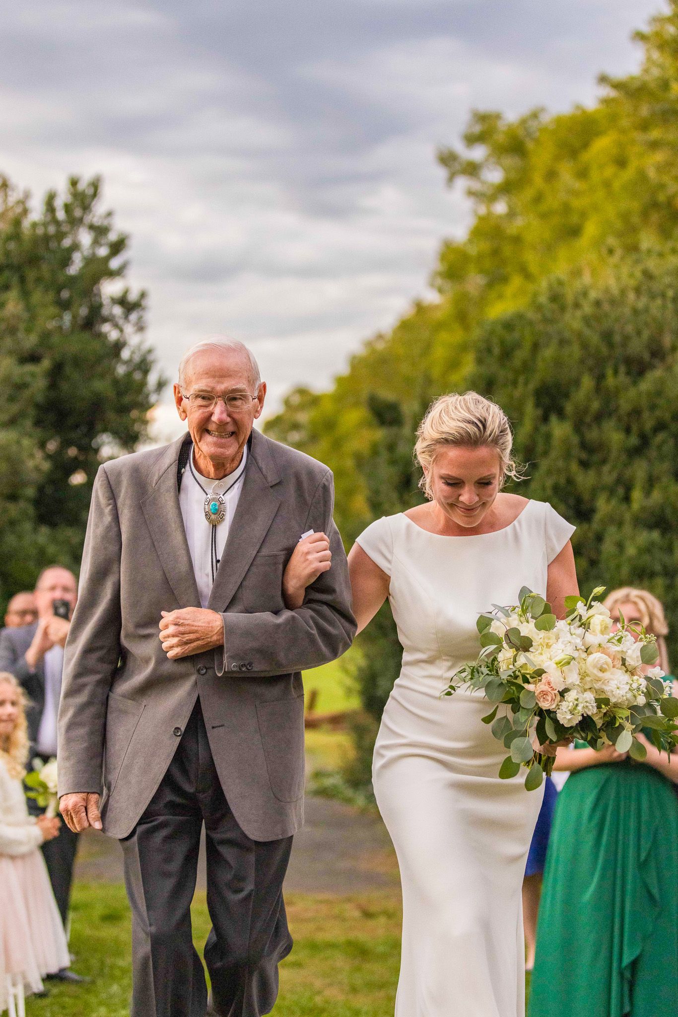 Wedding party members laugh together while walking outdoors on a cloudy day with greenery in the background.
