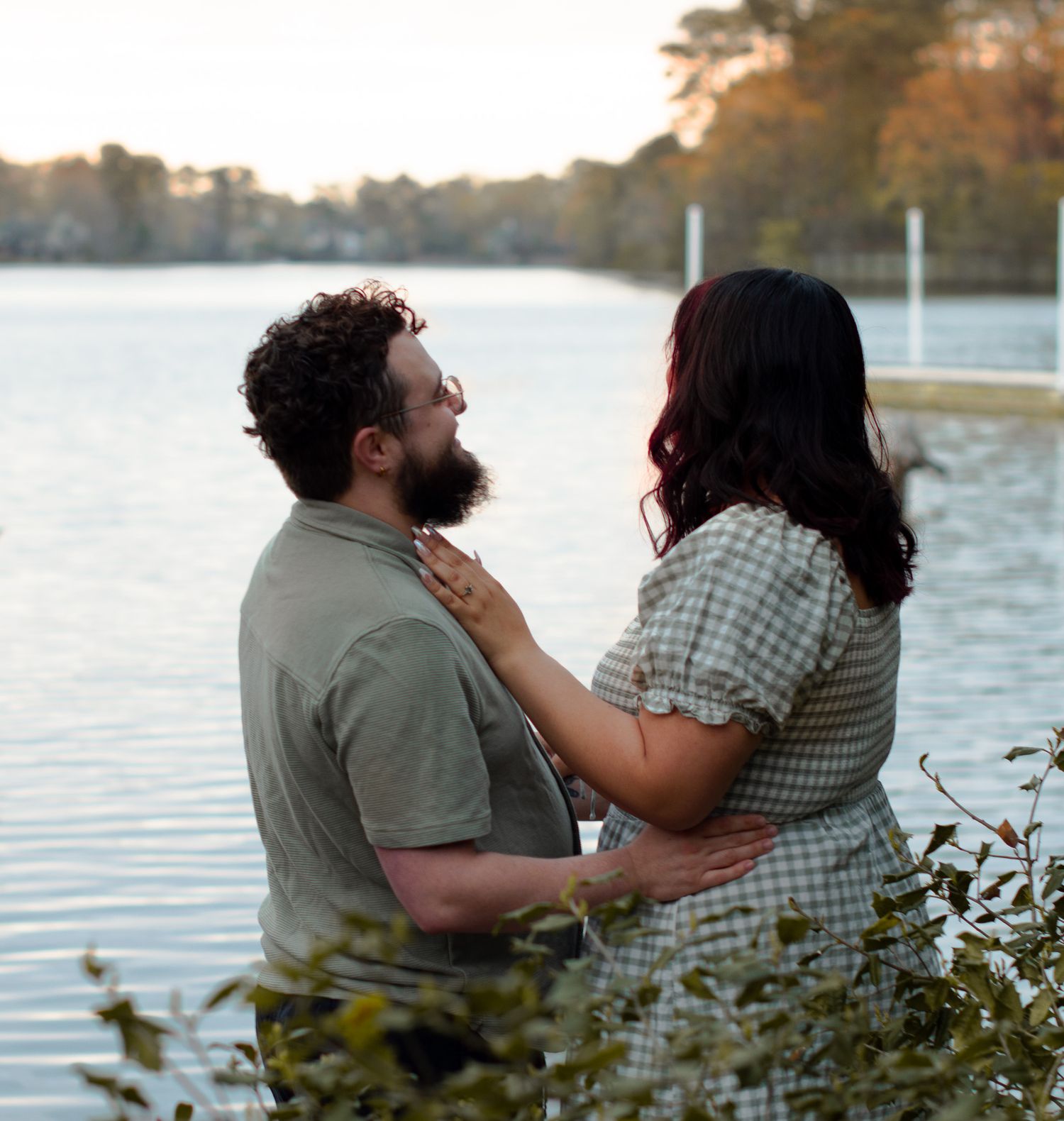Couple sharing a romantic moment by a serene lake at sunset, woman in plaid dress touching man's beard.