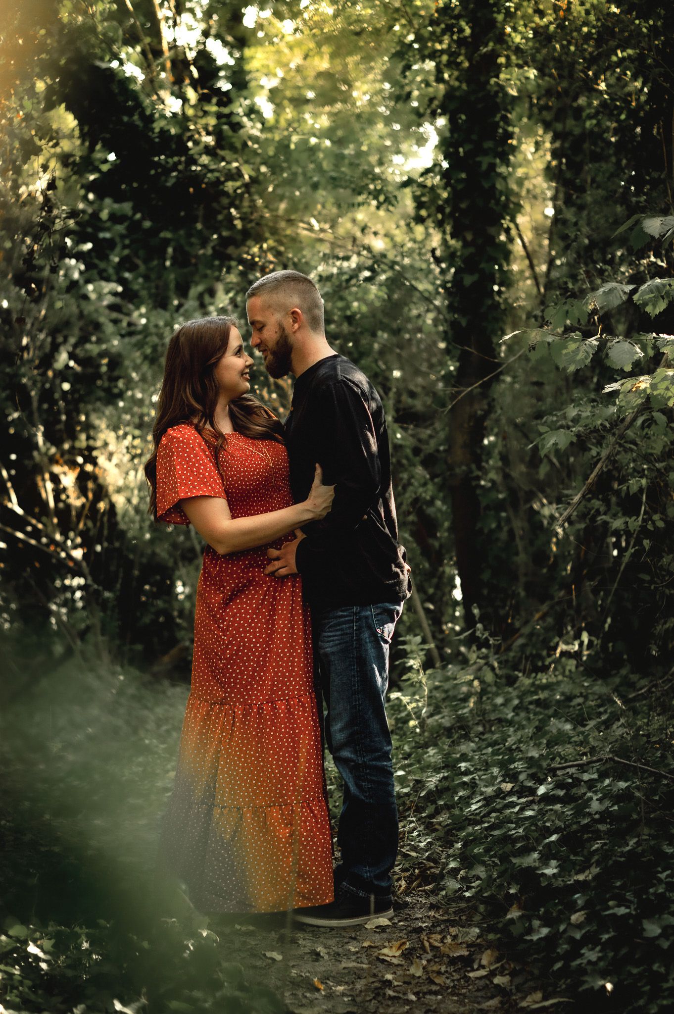 Romantic couple embracing in a sunlit wooded area with green foliage.
