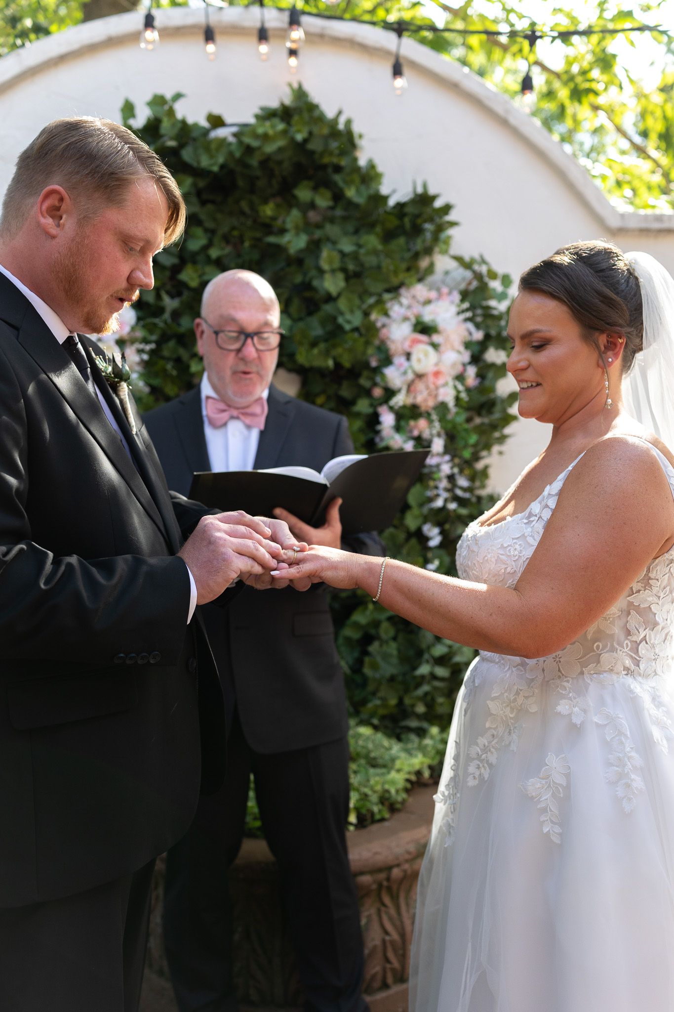 Groom places wedding ring on bride's finger during outdoor ceremony with officiant watching behind them.