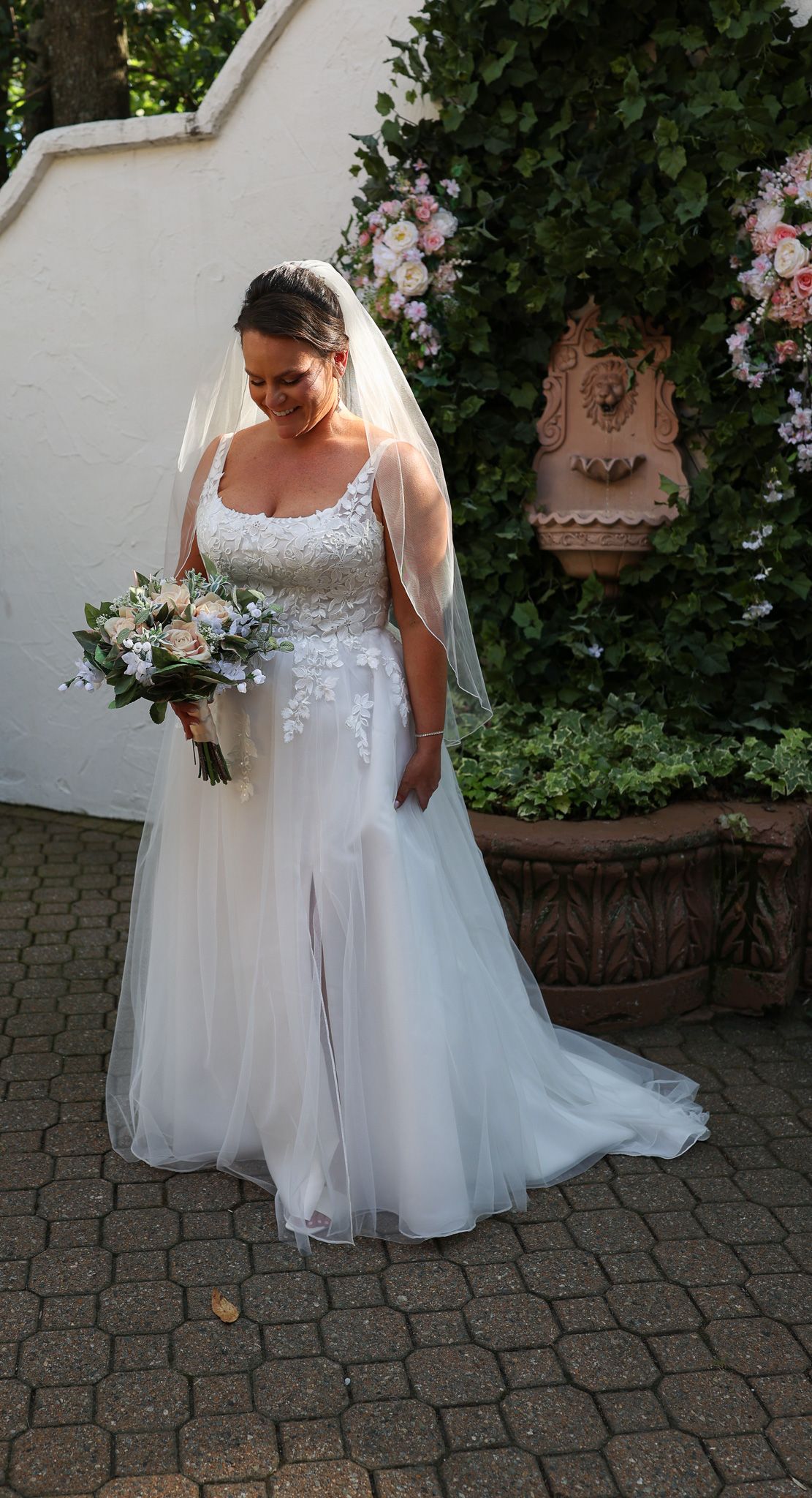Bride in a white lace A-line gown with veil holds a bouquet of roses in a garden setting.