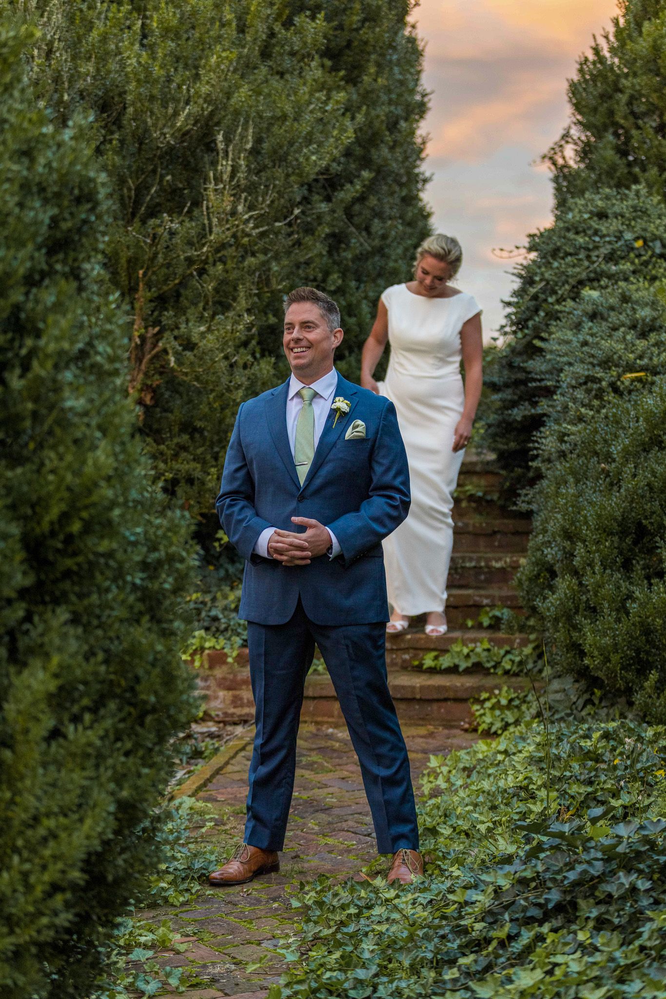 Person in navy blue suit stands in lush garden setting during sunset with greenery and stone walls in background.