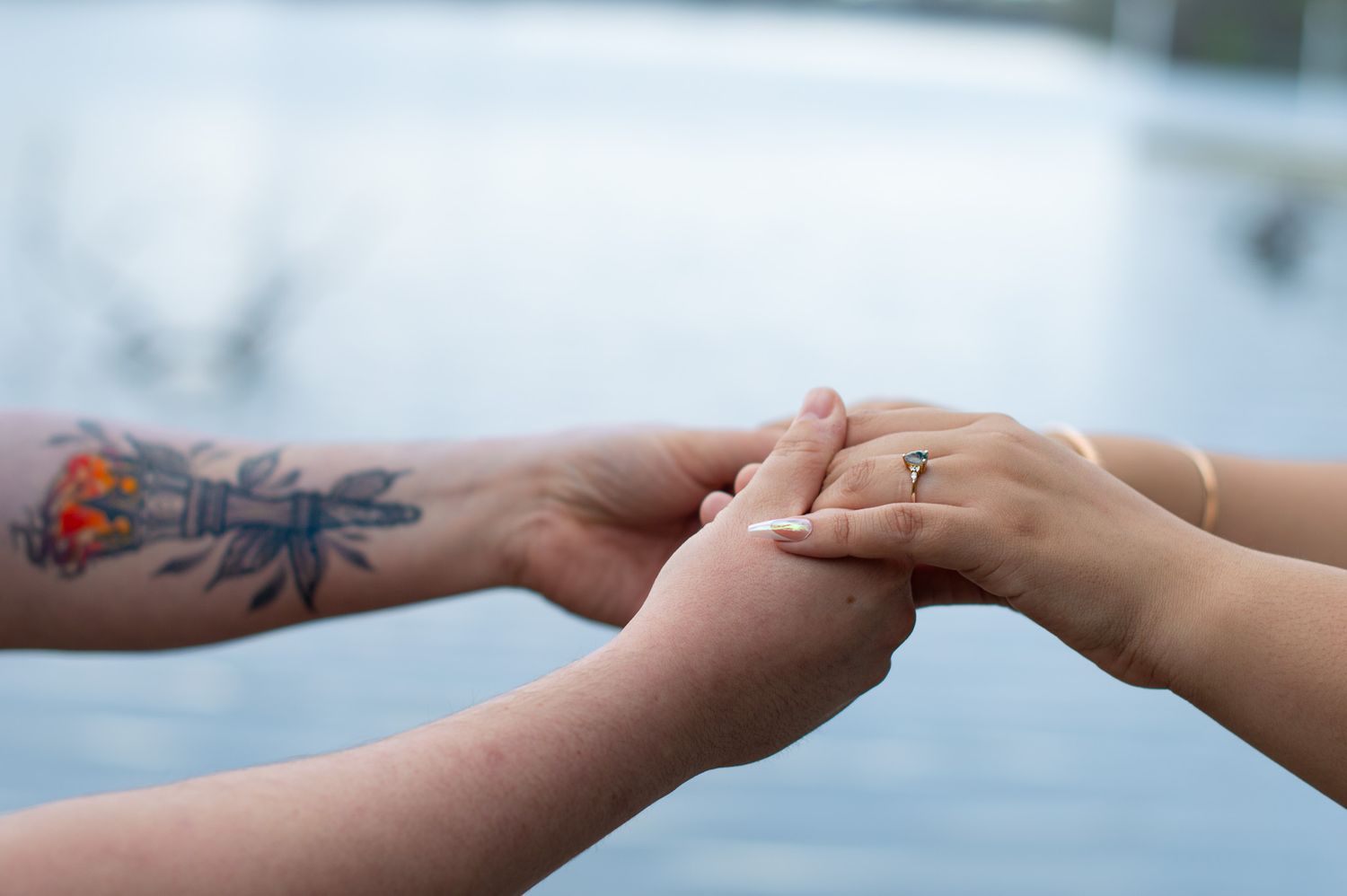 Two people holding hands, one with a colorful tattoo on their forearm and the other wearing a diamond ring.