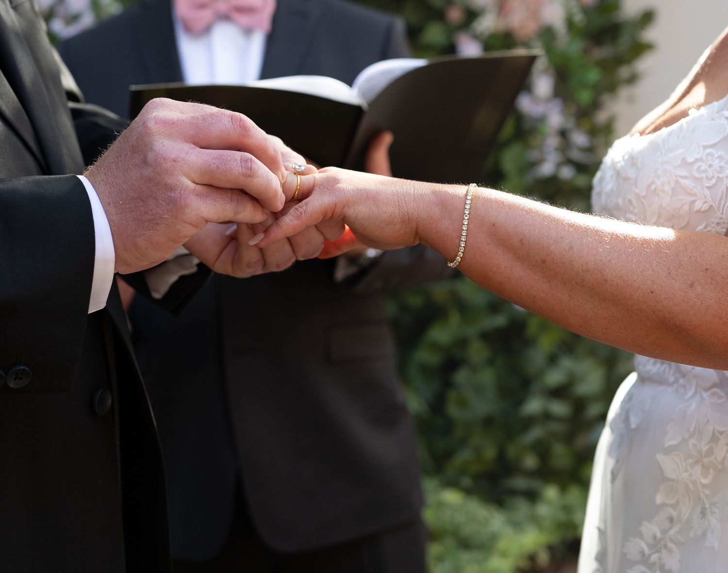 Groom placing a wedding ring on bride's finger during outdoor ceremony with officiant holding book in background.