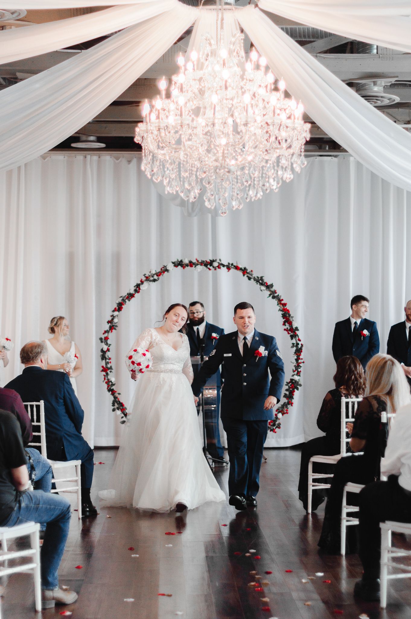 Elegant wedding ceremony setup with crystal chandeliers and floral arch in a white draped venue.