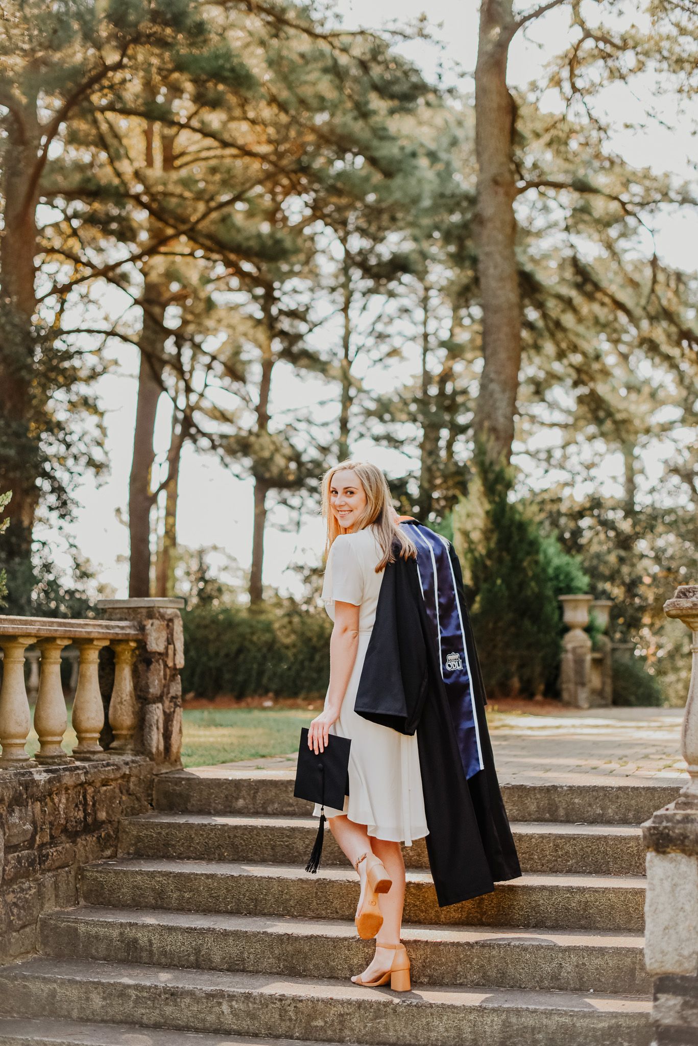 Graduate in cap and gown poses on stone steps surrounded by pine trees.