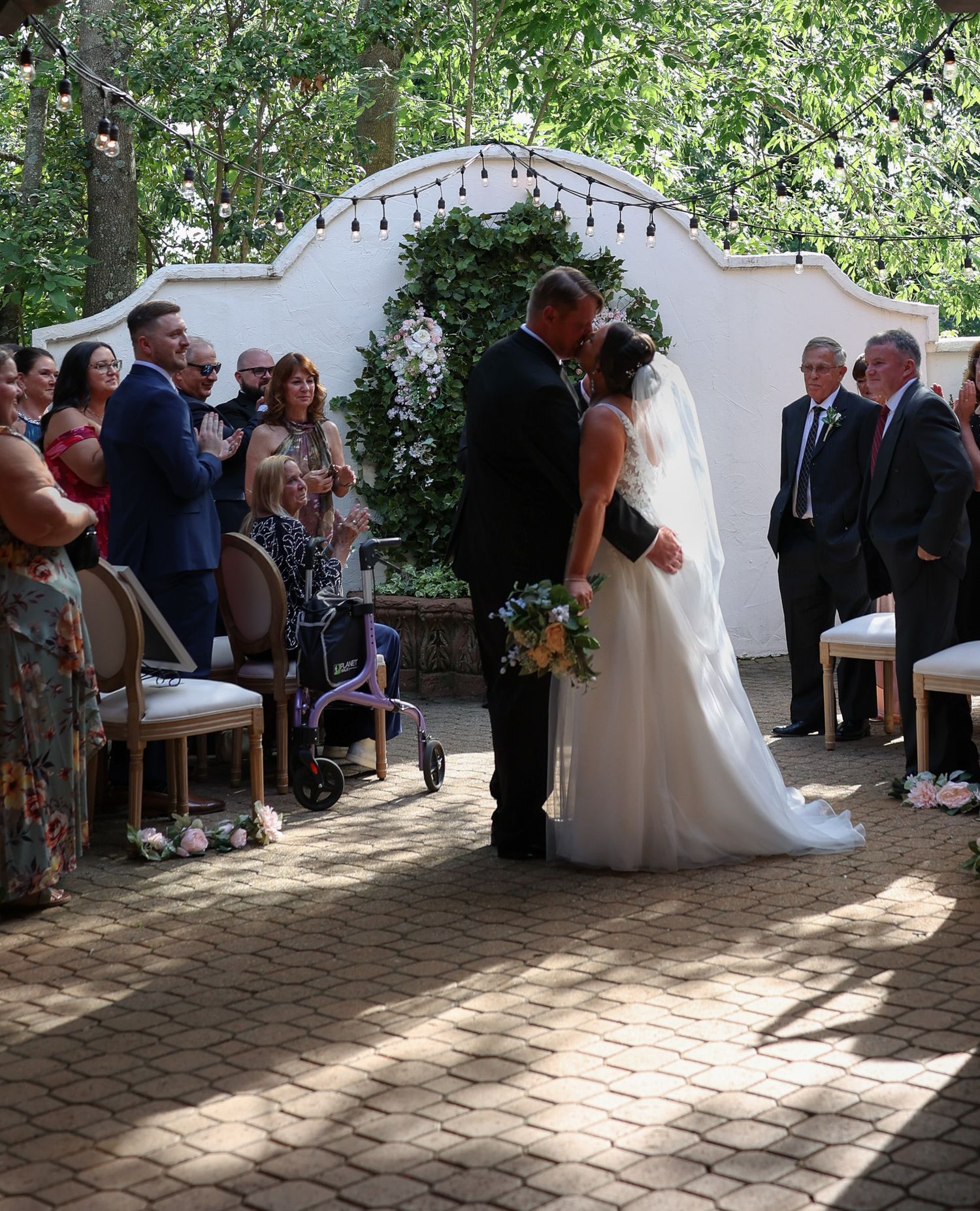 Bride and groom share first kiss at outdoor wedding ceremony surrounded by seated and standing guests.