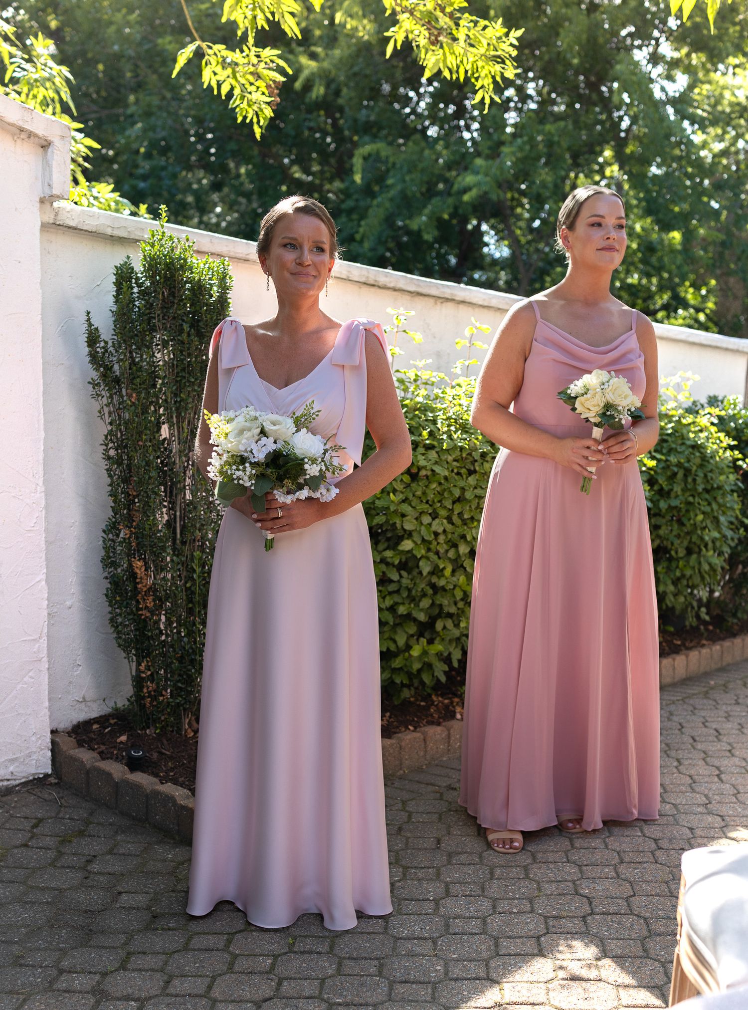 Two bridesmaids in pink gowns holding white bouquets stand outdoors at a wedding ceremony.