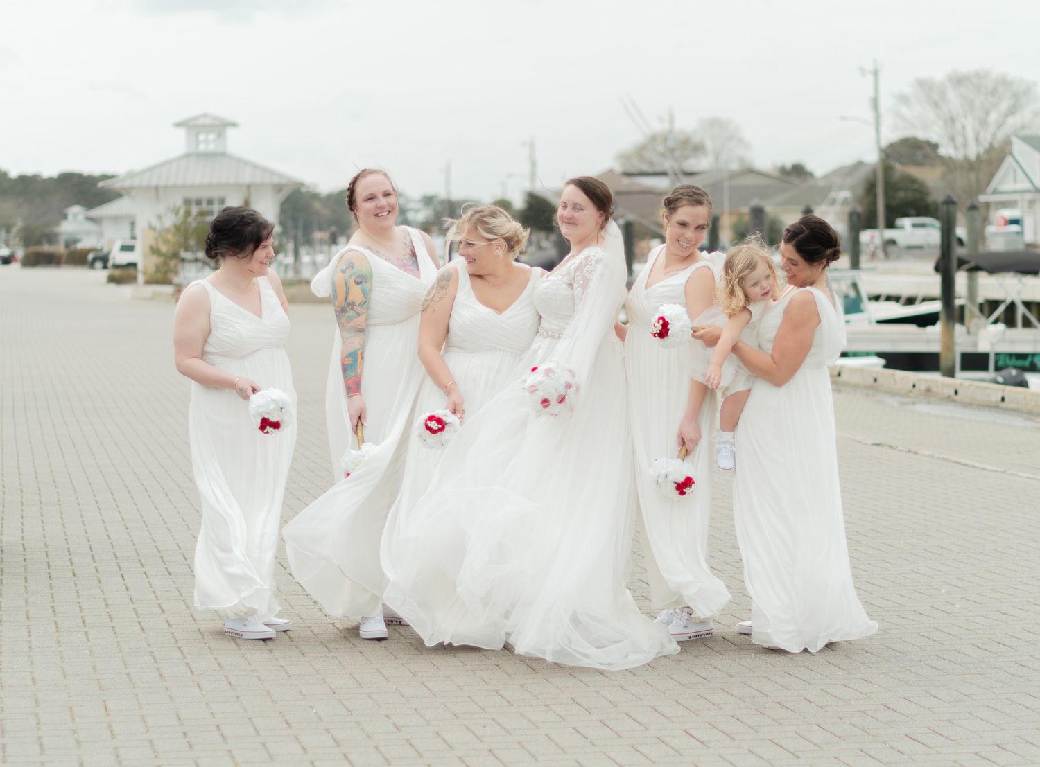 Bridesmaids in white dresses pose together on a paved outdoor area.