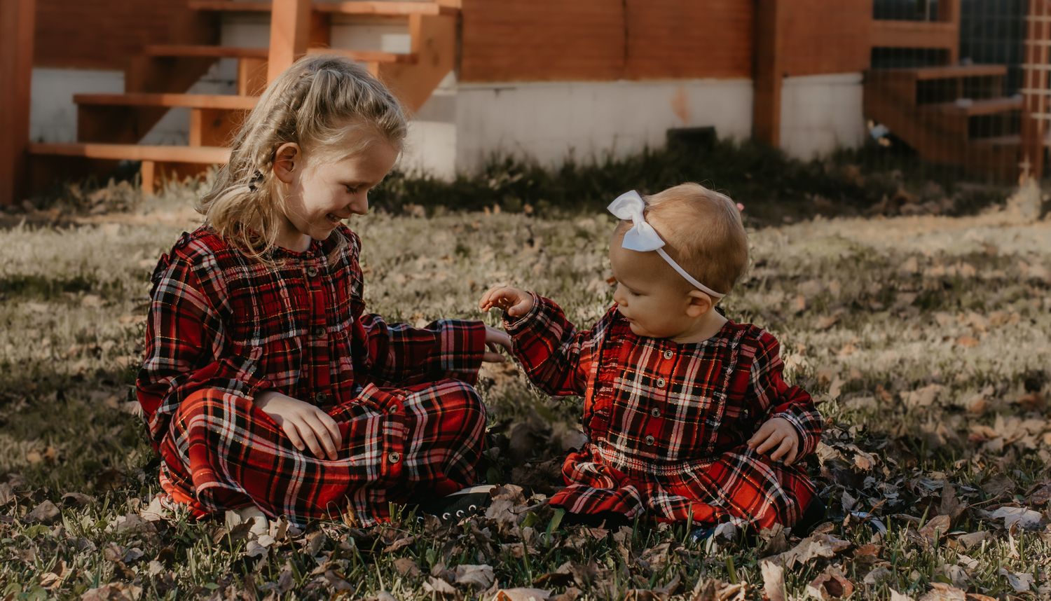 Two children in matching red plaid dresses sit together on grass outside a building.