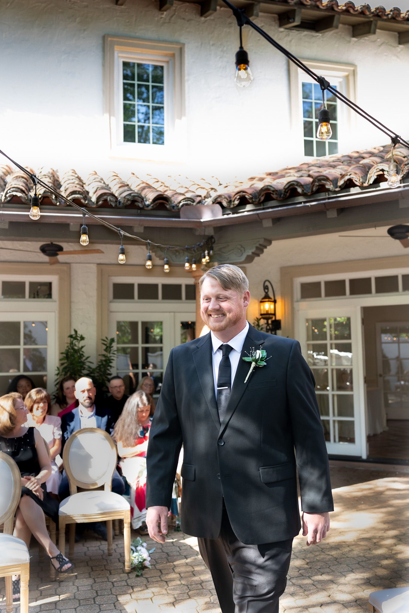 Smiling groom in black suit with boutonniere walks down outdoor wedding aisle at elegant venue with terracotta roof.
