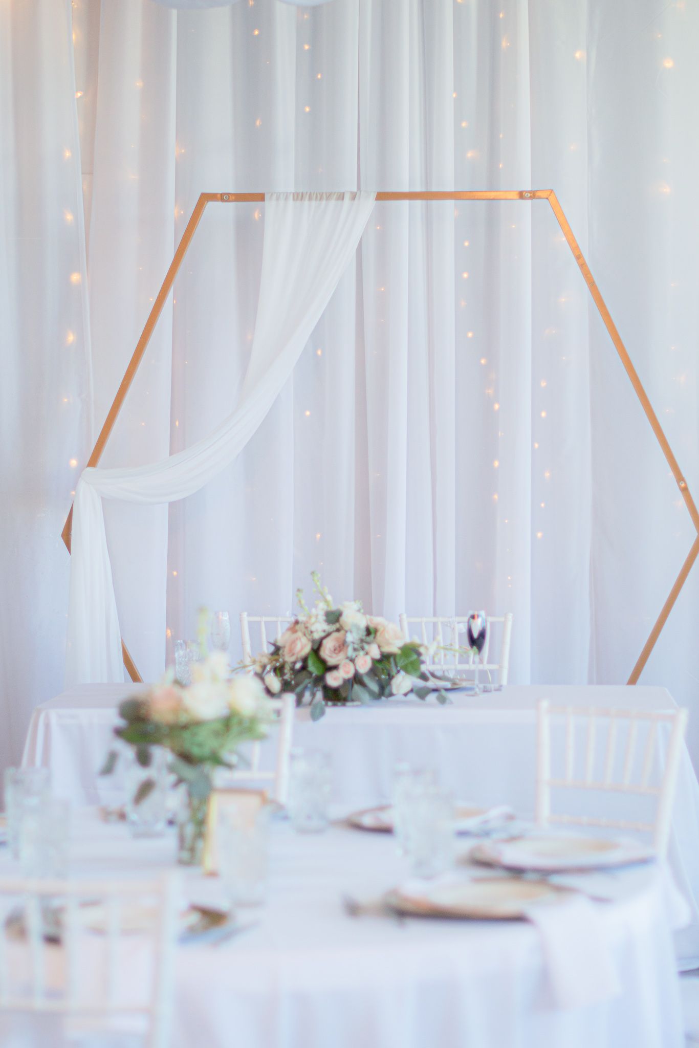 Elegant wedding reception table setup with hexagonal copper arch, white drapes, and delicate floral centerpieces.