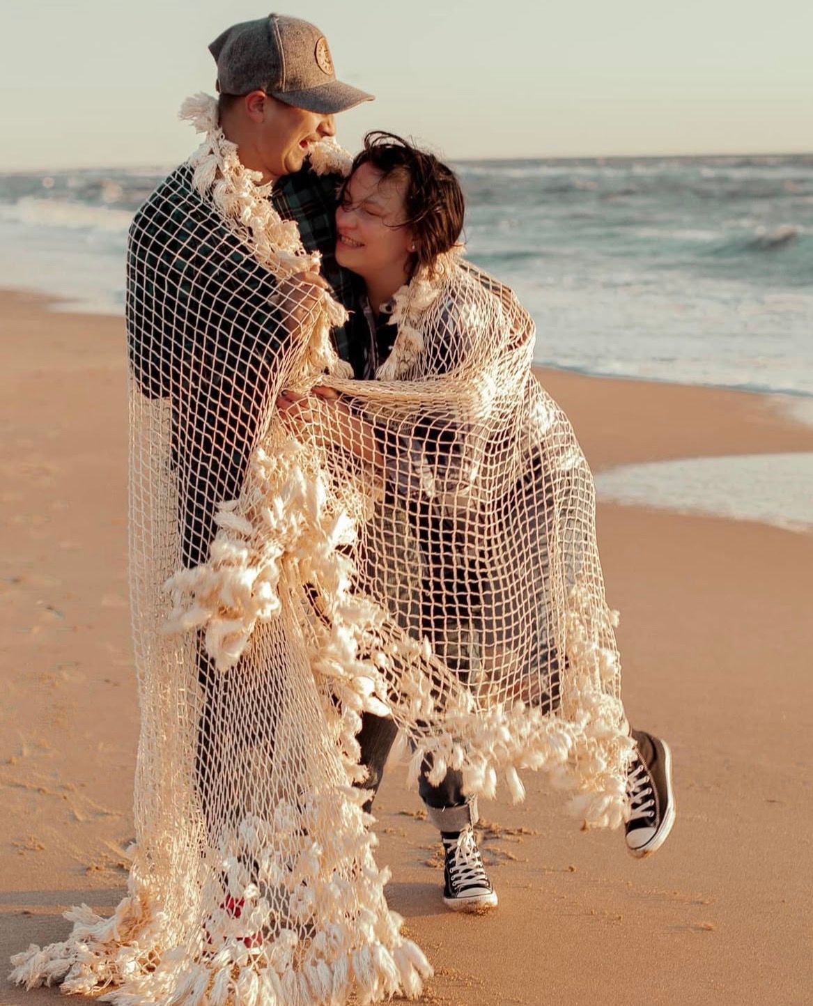 A couple wrapped in a delicate fishnet fabric embrace on a sunlit beach at golden hour.