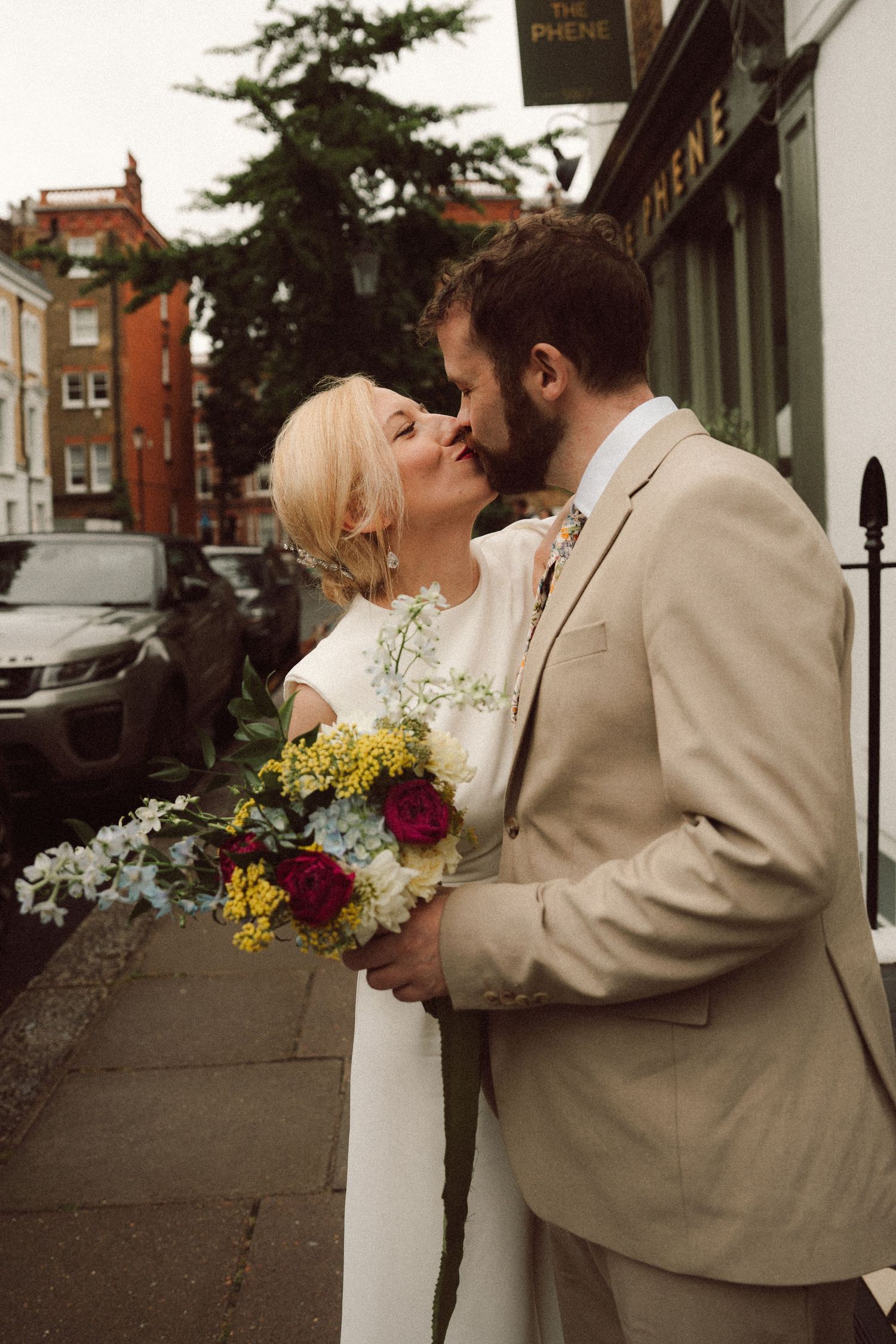 Bride and groom sharing a kiss at Chelsea Old Town H, bride holding a colourful bouquet of yellow, red, and white flowers.