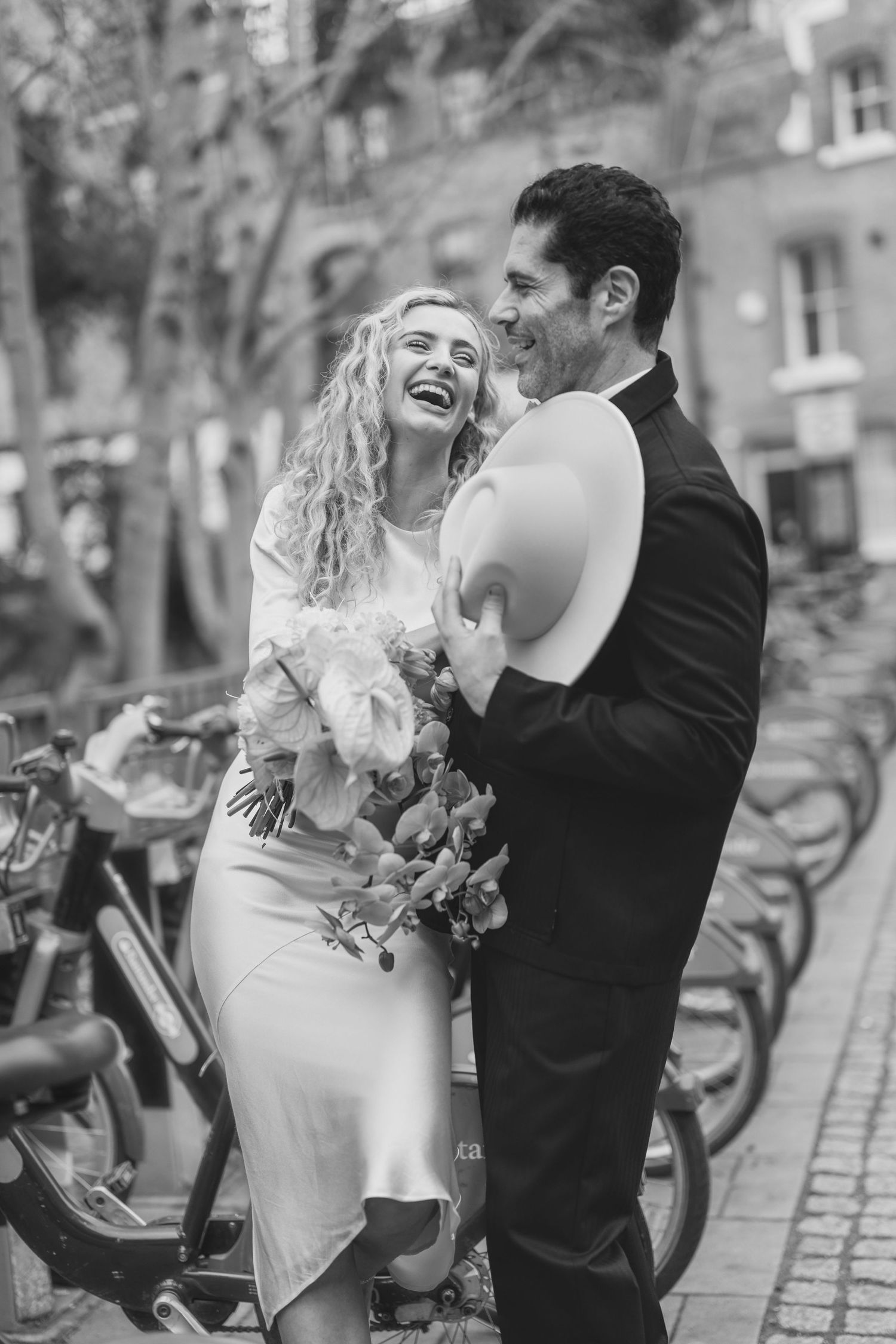 Black and white photo of a laughing bride holding flowers embracing a groom in a suit on a city street in London elopement wedding.