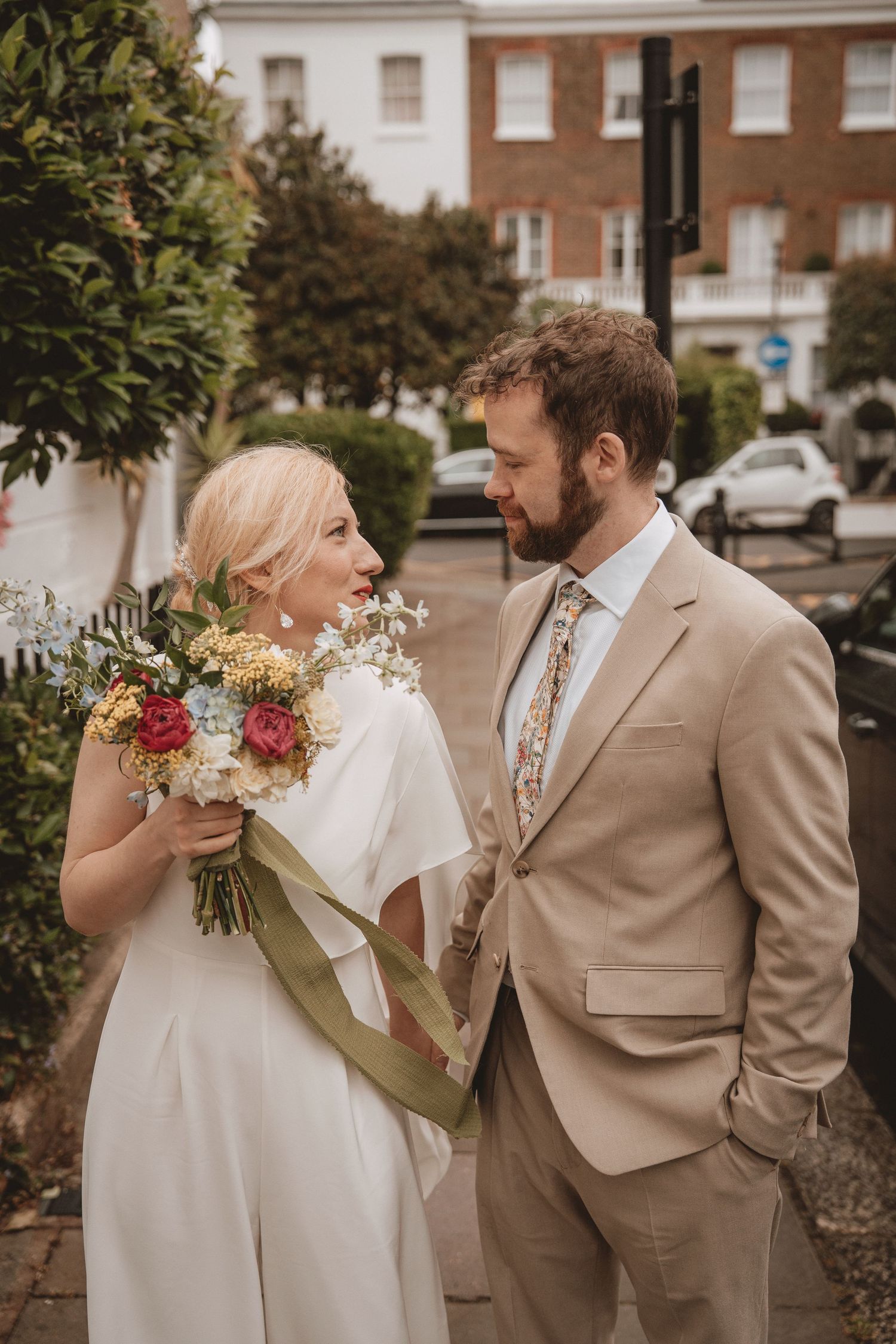 Bride holding colorful bouquet with green ribbon stands beside groom in tan suit on a tree-lined street at Chelsea Old Town Hall