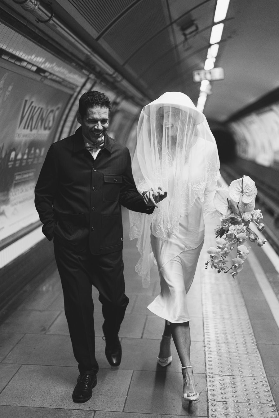 Black and white photo of a bride in veil and groom walking through a London Underground station platform.